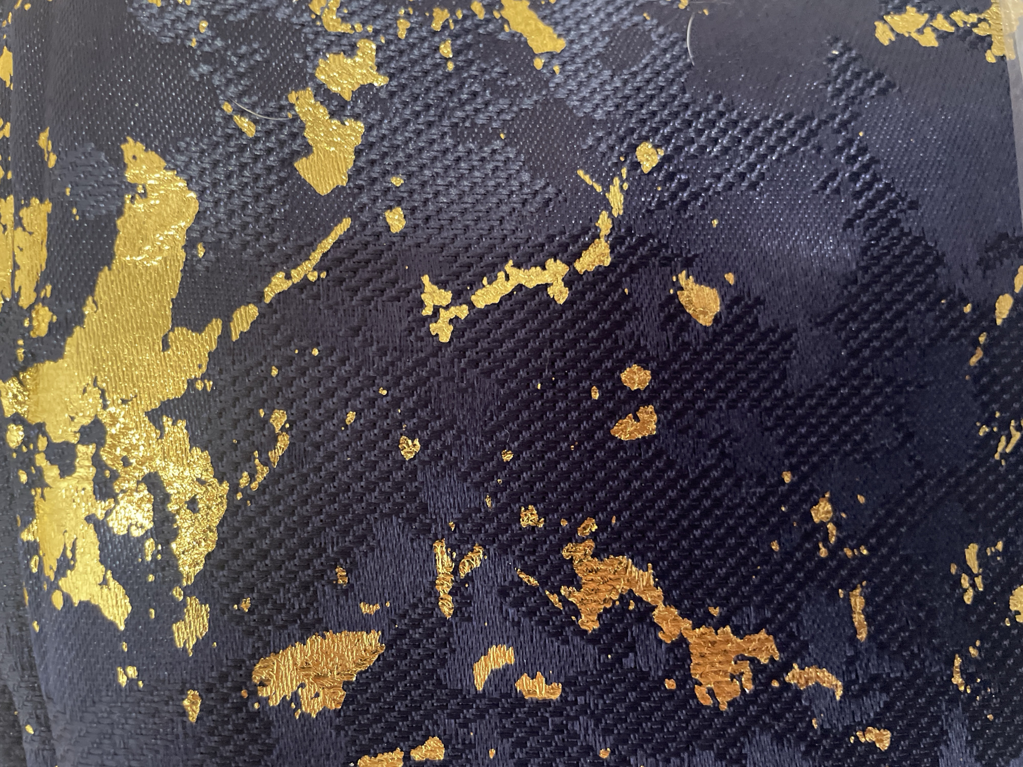 The image is a close-up, overhead shot of a dark blue textured fabric with a speckled gold pattern. The fabric appears to be some kind of upholstery or decorative material, possibly a velvet or a textured weave with a metallic overlay. The gold elements are irregular in shape and size, resembling splatters or abstract brushstrokes, giving the impression of a distressed or artistic finish. The texture of the dark blue fabric is visible, showing a weave pattern with small raised dots or threads, adding depth and dimension. The lighting highlights the metallic sheen of the gold, making it stand out against the dark background. The image is purely an abstract close-up of the material's pattern and texture. There are no people, objects, scenes, or identifiable settings visible. No activity or event is taking place. There is no visible text. The location context of Moscow, Russia, does not provide any visual cues within the image itself.