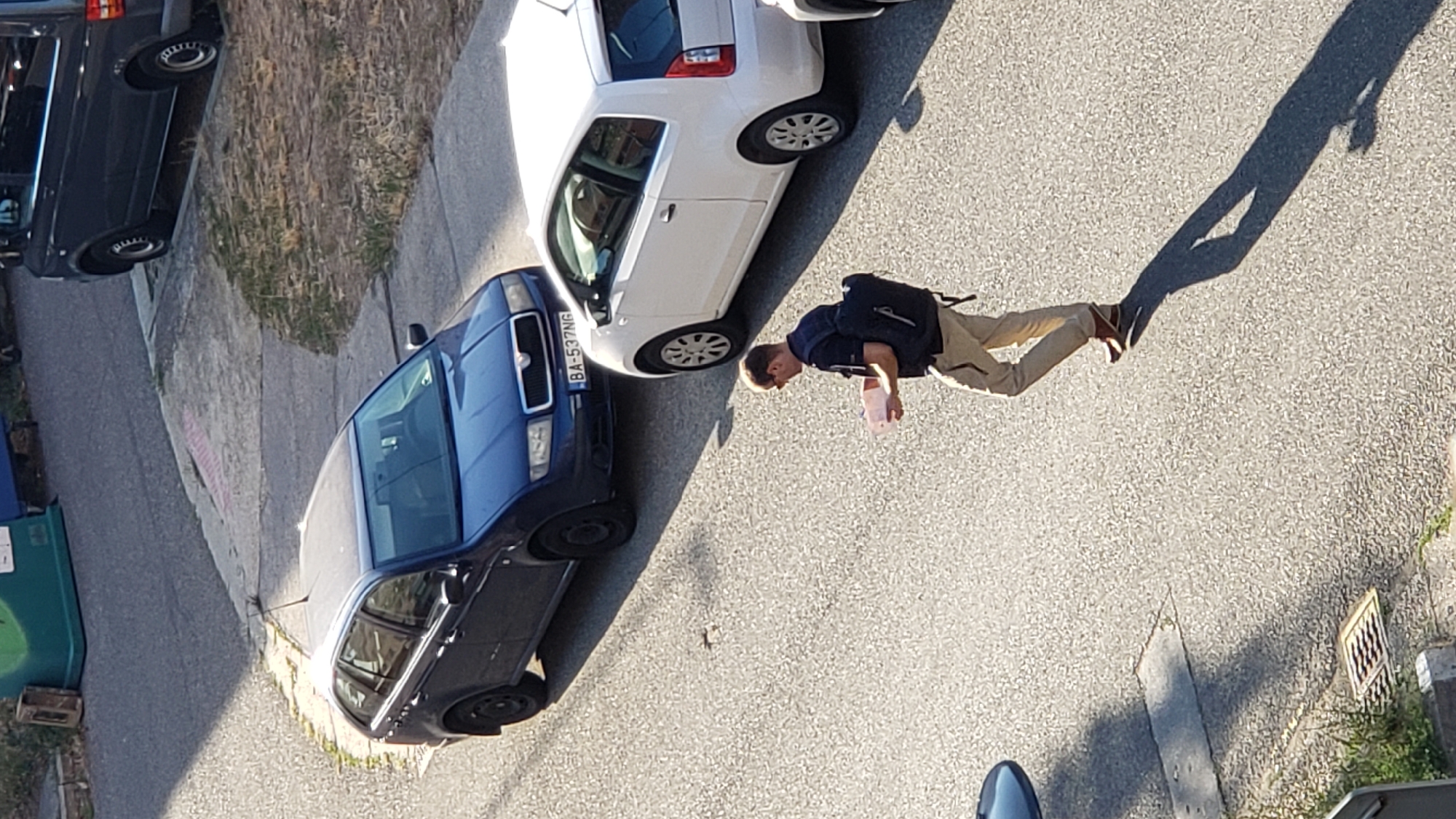 A man wearing a dark t-shirt, tan pants, and brown shoes is walking across a paved road, casting a long shadow. He has a black backpack on and is holding a light-colored piece of paper or small box in his hands. He is walking away from the camera, towards the right side of the frame.
In the background, two cars are parked on the road. The car on the left is a dark blue station wagon with a license plate that reads "BA-537NG". The car on the right is a white hatchback. Further in the background, a dark colored van is parked, and behind that, several large green and blue bins are visible. The scene appears to be in a residential area, possibly a street or parking lot, with some dry grass and patchy vegetation visible along the edges. The light suggests it is daytime, likely late afternoon due to the long shadows.
The license plate indicates the location as Bratislava, Slovakia.