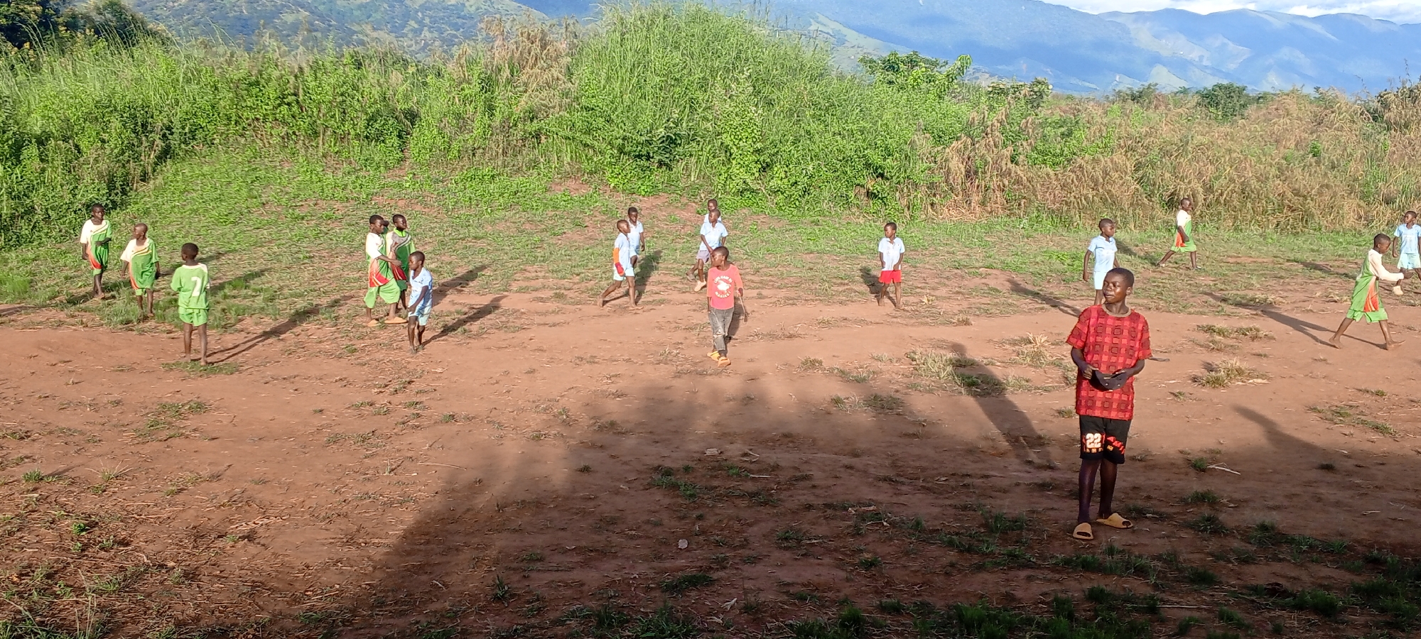 The image depicts a group of children in Ngalula, Democratic Republic of the Congo, playing outdoors. The setting is a dry, grassy field with some vegetation. In the background, there are hills or small mountains. The sky is visible in the background. There are approximately 15 children of varying ages. They are dressed in a mix of clothing, including green jerseys, white shirts, blue shirts, red shirts, and colorful patterns. Some are barefoot, while others wear sandals. Most of the children are scattered across the field, seemingly engaged in some sort of game or activity. One child, wearing a red checkered shirt, black shorts with the number 22 on them, and yellow sandals, stands in the foreground, looking towards the camera's right side. His shadow is cast prominently on the ground.