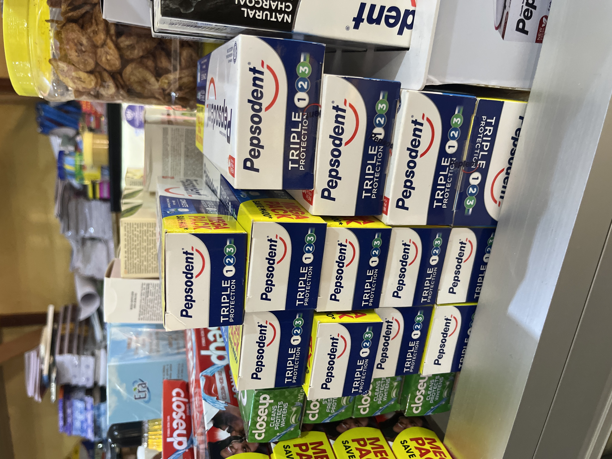 The image shows a display of Pepsodent toothpaste boxes and Close Up toothpaste boxes stacked on a shelf in a store, likely a local shop or market stall. The Pepsodent boxes are predominantly white with blue and red accents, featuring the brand logo and the text "TRIPLE PROTECTION 1 2 3". Some boxes are oriented vertically, while others are stacked horizontally. The Close Up boxes are green with white text. In the background, there are other products, including what appears to be a jar of dried banana chips and some stacked paper items. A red and white box with the brand "Close Up" and an image of a smiling person is partially visible on the left. The overall impression is of a retail environment stocked with personal hygiene products. The image is taken from a slightly elevated angle, looking down at the products. The lighting suggests an indoor setting with ambient light. The location is identified as Bukuru, Nigeria.