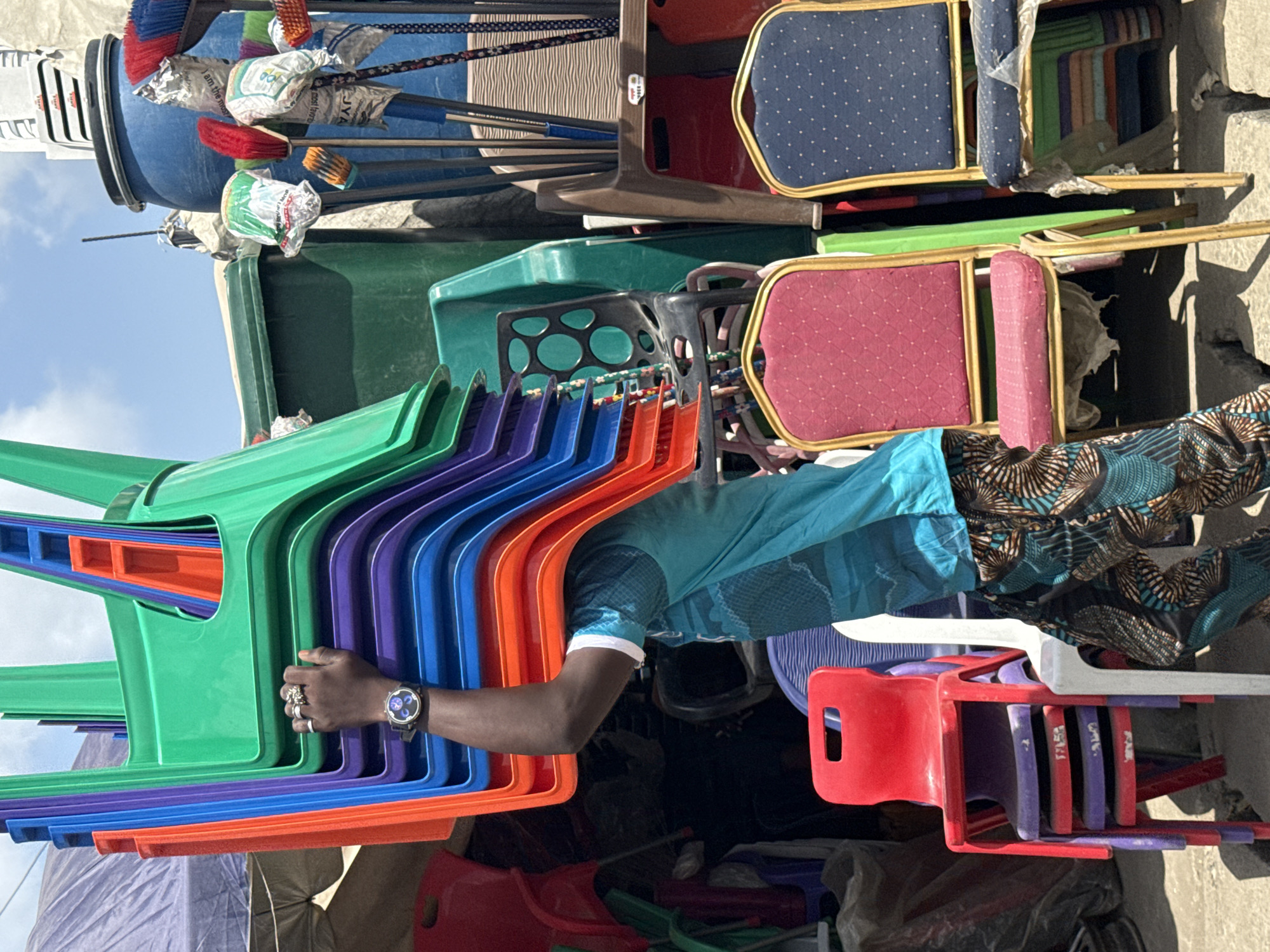 In Ado, Nigeria, the scene depicts a person wearing a blue shirt, printed fabric, and a wristwatch, holding a stack of colorful plastic chairs (green, purple, blue, and orange). These chairs are stacked next to upholstered chairs (blue, red, and green) with gold trim and a pile of brooms. The background consists of bins and other objects, suggesting a storage or sales area.