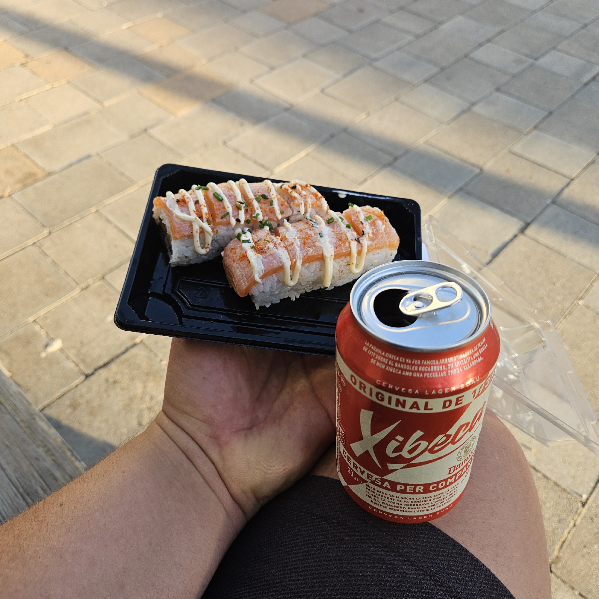 A person's arm and hand hold a black plastic container of sushi rolls topped with white sauce and chives, next to a red and white can of Xibèrca lager beer. The sushi and beer are placed on a light-colored tiled surface, casting shadows from an unseen light source. The scene appears to be outdoors, possibly a public space in El Perelló, Spain, given the branding on the beer can. The person is wearing dark clothing.