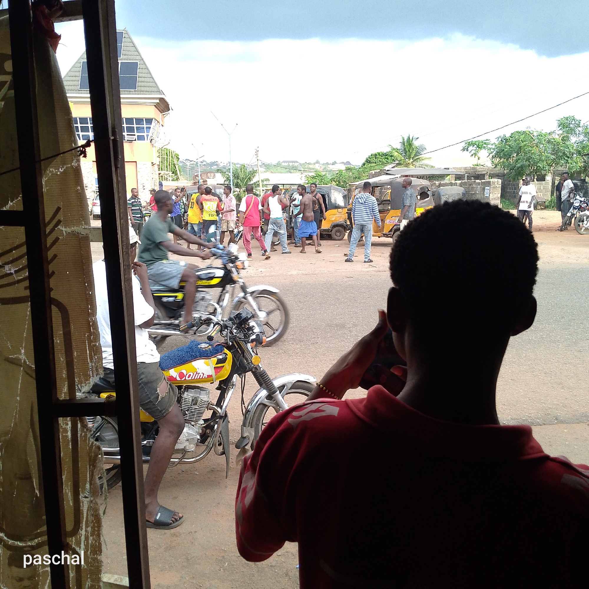 The image shows a street scene in Nsugpe, Nigeria, viewed through a damaged window or doorway. Several people are gathered in the street, some of whom appear to be interacting or possibly arguing. Two motorcycles are visible in the foreground, one occupied. A three-wheeled vehicle (likely a "keke" or auto-rickshaw) is parked nearby. A building with solar panels is seen in the background. The individual in the foreground is possibly talking on the phone.