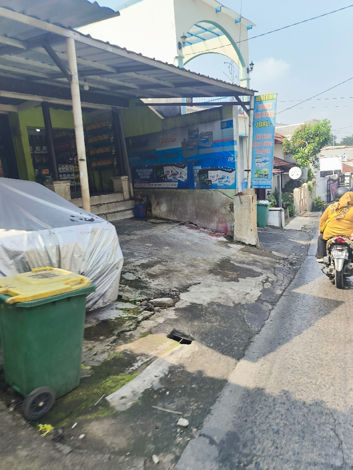 The image depicts a narrow street in Bandung, Indonesia (latitude: -6.909, longitude: 107.704) with a car covered in a gray tarp parked on the left side of the street. A green trashcan with a yellow lid sits in front of the car. A person is riding a motorcycle on the right side of the street. There is a blue sign advertising batteries on the side of the building on the left side of the street. The street is made of concrete and appears to be in a state of disrepair, with cracks and patches. There are green plants growing in the cracks in the concrete. The building on the left side of the street has a faded green exterior and a gray awning. The building has a metal roof.
