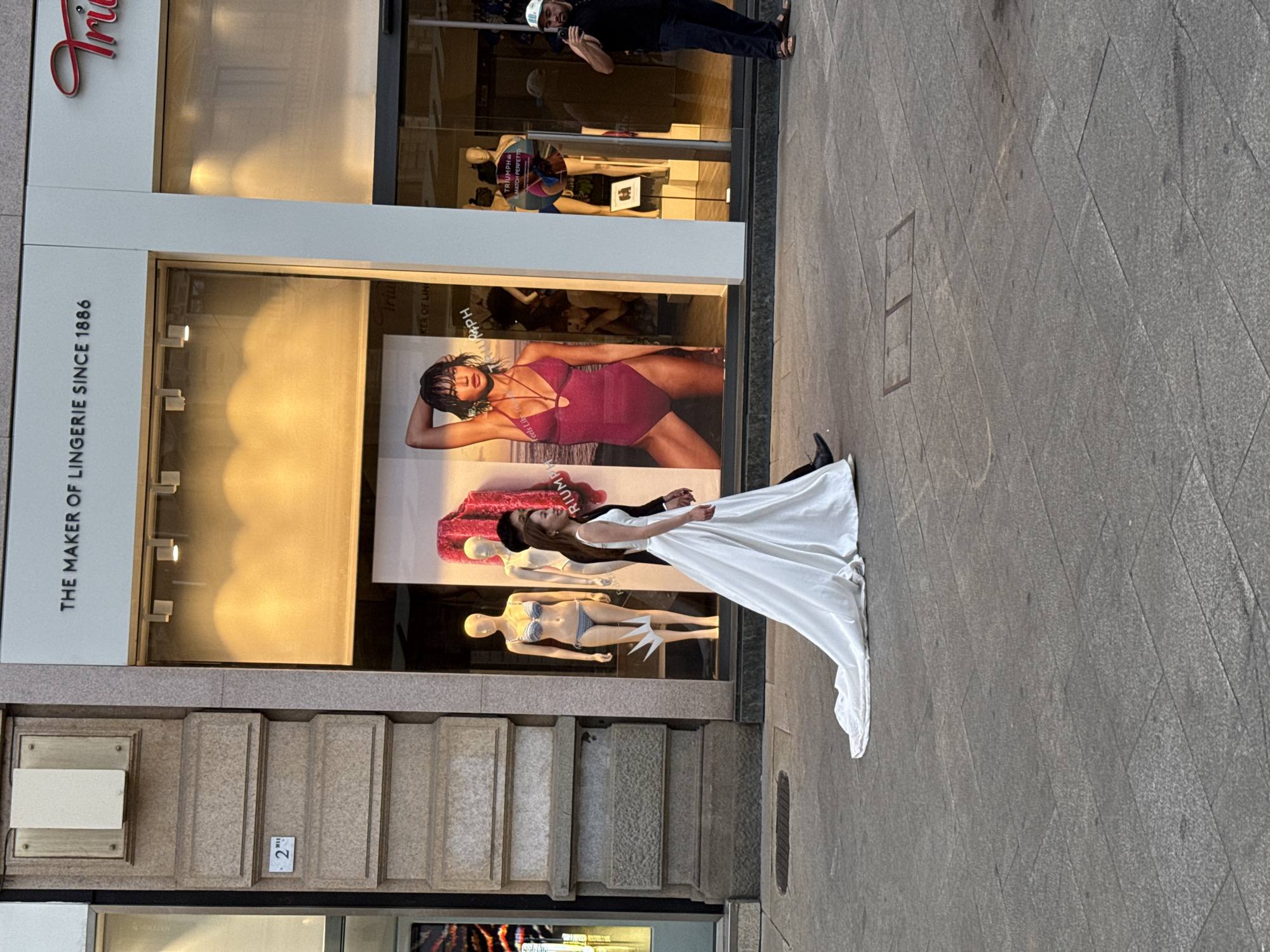 A young woman in a white wedding dress with a long train is walking on a paved street in Milan, Italy. She is accompanied by a man in a dark suit. They are walking in front of a storefront with a large advertisement featuring a woman in a red swimsuit. The storefront also displays mannequins wearing lingerie. The street is made of large granite slabs and has metal grates embedded in it. The buildings surrounding the street are made of stone. The sky is not visible.