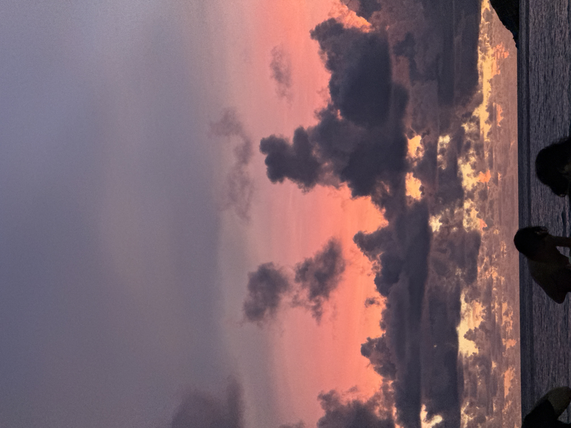 This is a photograph of a dramatic sunset over the ocean. The sky is filled with vibrant colors, ranging from deep purples and grays to warm oranges and pinks. Dark, heavy clouds are contrasted with brighter, illuminated patches of sky, creating a sense of depth and intensity. The sun appears to be setting on the right side of the frame, casting a golden glow on the clouds and the water.
On the right side of the image, silhouetted figures of people are visible, suggesting they are either standing on a beach or a pier, enjoying the sunset. The water stretches out towards the horizon, reflecting the colors of the sky. The overall scene evokes a sense of awe and tranquility, characteristic of a beautiful sunset.
The location is identified as Rawai, Thailand, which is known for its beautiful beaches and ocean views, making it a popular spot for sunset viewing. The time of day is clearly sunset, indicated by the warm, low-angled light and the colors in the sky. The weather appears to be clear enough for a sunset, despite the presence of dramatic cloud formations. There is no visible text in the image.