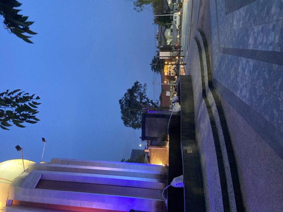 The image shows a view of a fountain in the city of Sidoarjo, Indonesia, at coordinates -7.446 latitude and 112.719 longitude. The fountain is illuminated with purple and blue lights, and there are two jets of water shooting upwards from the base. The fountain is situated in a paved area, with steps leading down to a lower level. The surrounding area is dark, with only a few streetlights illuminating the scene. There are trees on the right side of the image, and a building in the background, illuminated with yellow light. In the distance, there are cars driving on a street with streetlights. The sky is a dark blue, with a few stars visible. The image was taken at dusk.