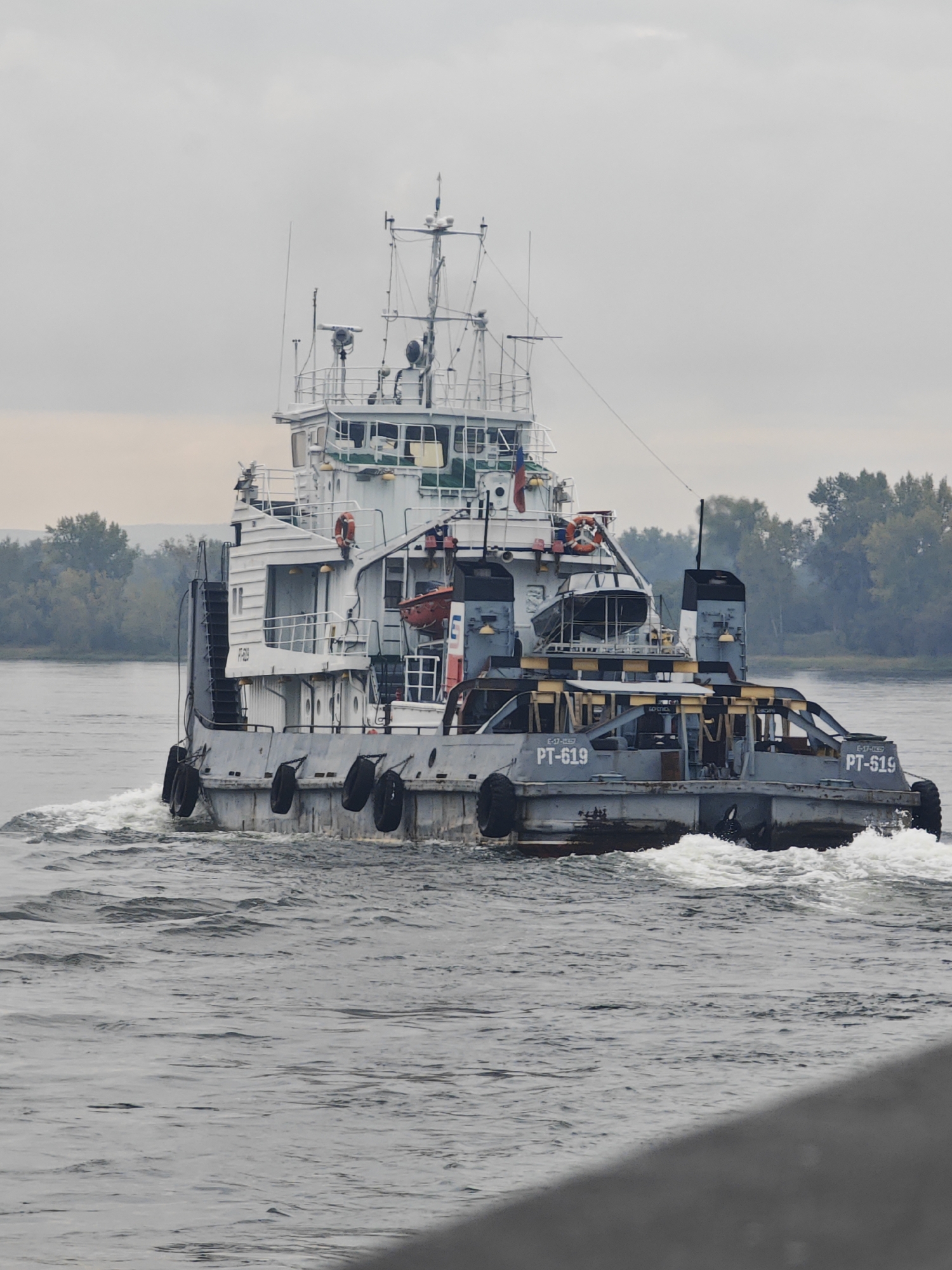A gray and white tugboat, identified by the markings "PT-619" and "PT-519," navigates a body of water. The tugboat is a multi-level vessel with a white superstructure and gray hull, equipped with various antennas and navigational equipment. It is creating a wake as it moves through the water. The background consists of a treeline with autumn foliage and a hazy, overcast sky. The image appears to be taken from a slightly elevated perspective, possibly from another boat or a dock, with a gray, sloped foreground element on the right side of the frame. The location is identified as Ермолаево, Russia.