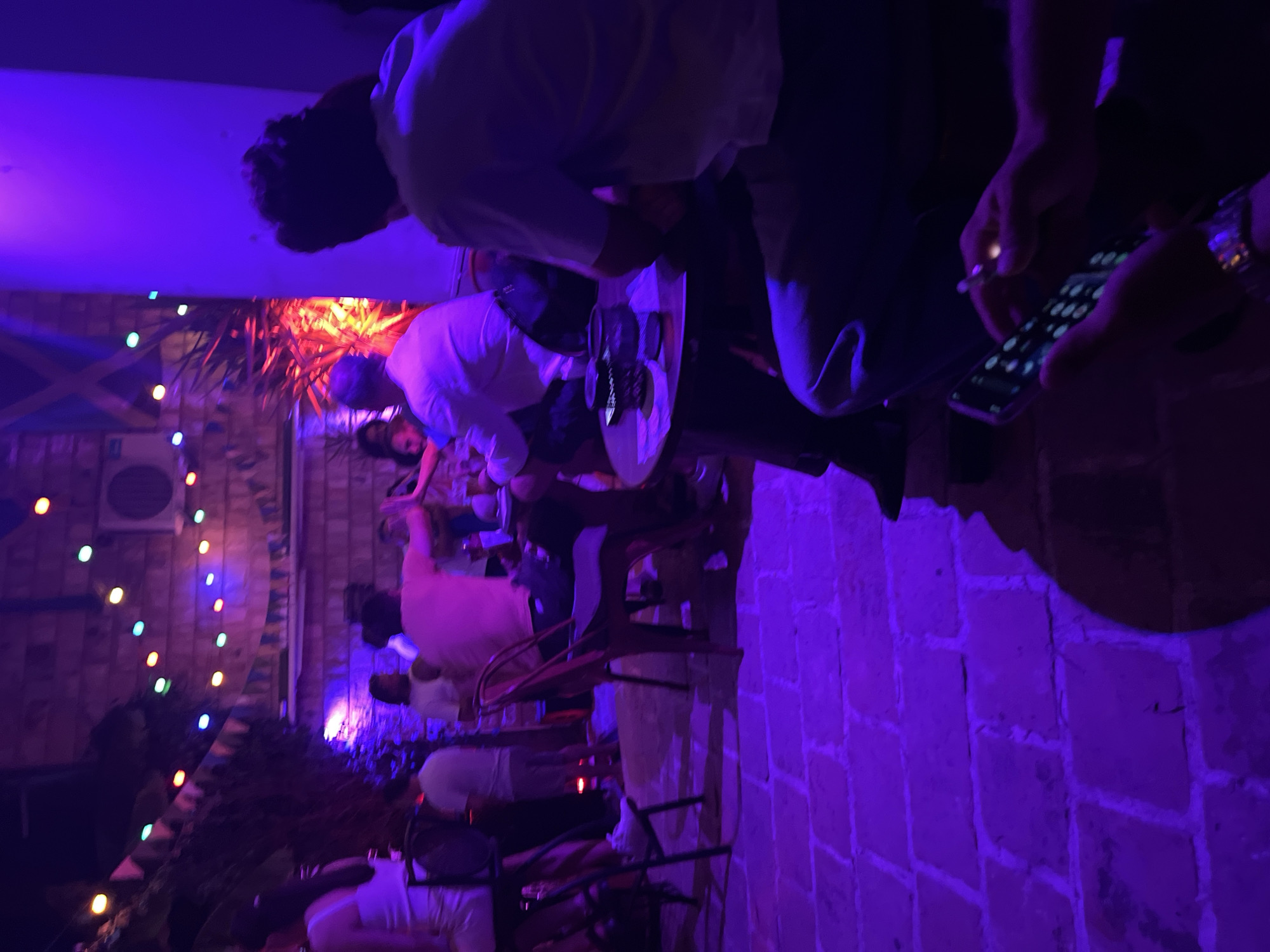 The image depicts an outdoor gathering at night, illuminated by colorful string lights and purple and red lighting, suggesting a bar or restaurant setting in Herceg Novi, Montenegro. Several individuals are present, some seated at tables, others standing. In the foreground, a person is holding a smartphone and what appears to be a cigarette. The ground is paved with stone tiles. The overall atmosphere is festive and social.