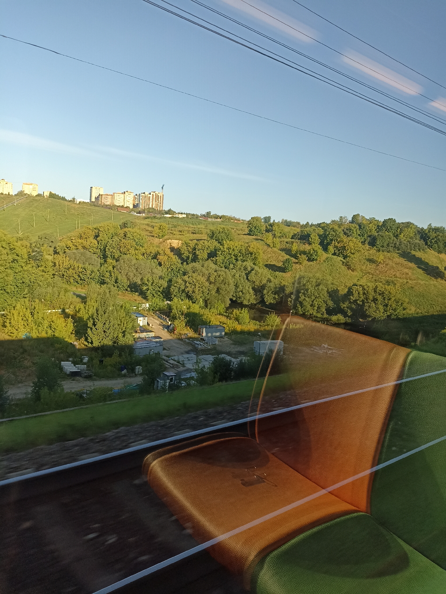 The image, taken from inside a moving train, shows a view of a suburban landscape under a clear blue sky. In the foreground, a train seat with orange and green upholstery is visible, partially reflecting the outside scenery. The window also shows reflections of overhead power lines.
The exterior view features a gently sloping, grassy hill dotted with trees. Several buildings, appearing to be apartment complexes, are situated on the crest of the hill. A construction crane is visible near one of the buildings. Below the hill, there is a small cluster of industrial or storage buildings, some with white roofs, and what appears to be a dirt road or pathway. Further down, there is a line of trees and green fields. The sun appears to be low in the sky, casting long shadows. The overall impression is that of a rural or semi-rural area on the outskirts of a city.