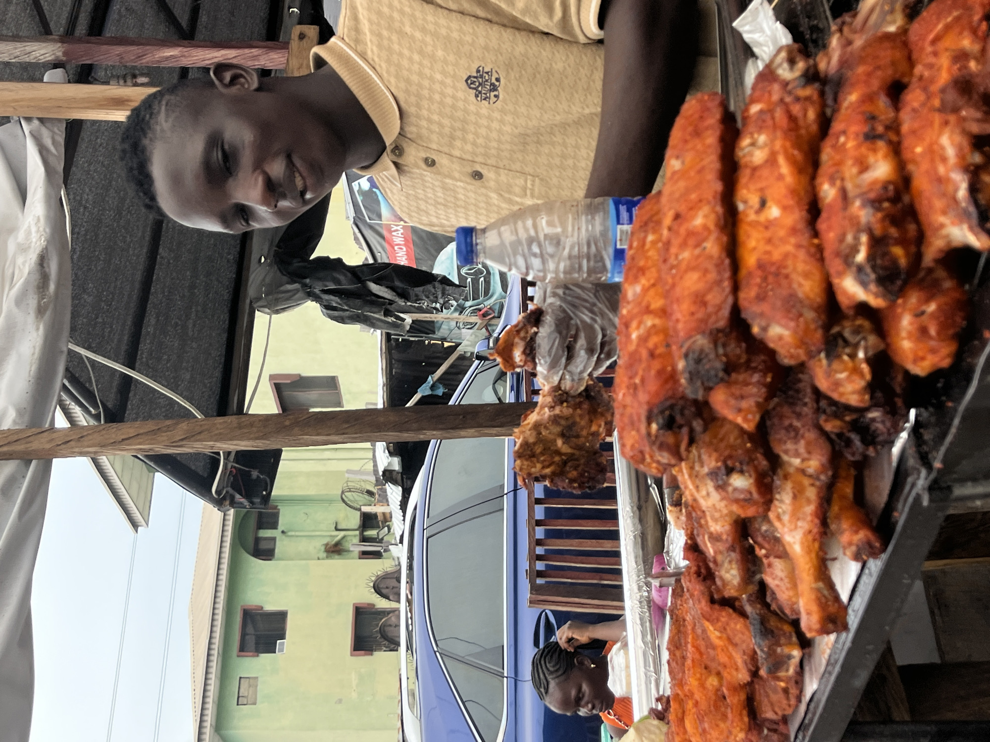 A man in a light yellow shirt stands next to a pile of cooked turkey in a tray. He is smiling at the camera. Behind him are a clear water bottle and a black car, and in the background, there is a light green building with windows. Another person stands to the right of the car. The image was taken in the city of Ado, Nigeria.