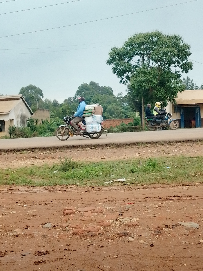 The image shows a man riding a motorcycle on a paved road in the town of Kivaywa, Kenya. The man is wearing a blue shirt and a hat, and the motorcycle is carrying a large load of packages. There is a second motorcycle with two riders in the distance, as well as a building and trees. The road is muddy and dusty. The image was taken from a point in the town of Kivaywa, Kenya, at the coordinates latitude: 0.56, longitude: 34.806. The scene appears to be on the outskirts of town, with a single-story building visible behind the motorcycles. The man on the motorcycle is heading towards the viewer. The motorcycles are traveling at a speed that is consistent with travel on a town road. It is a quiet scene, and the only sound is the hum of the engines. The weather is overcast, but the air is still.