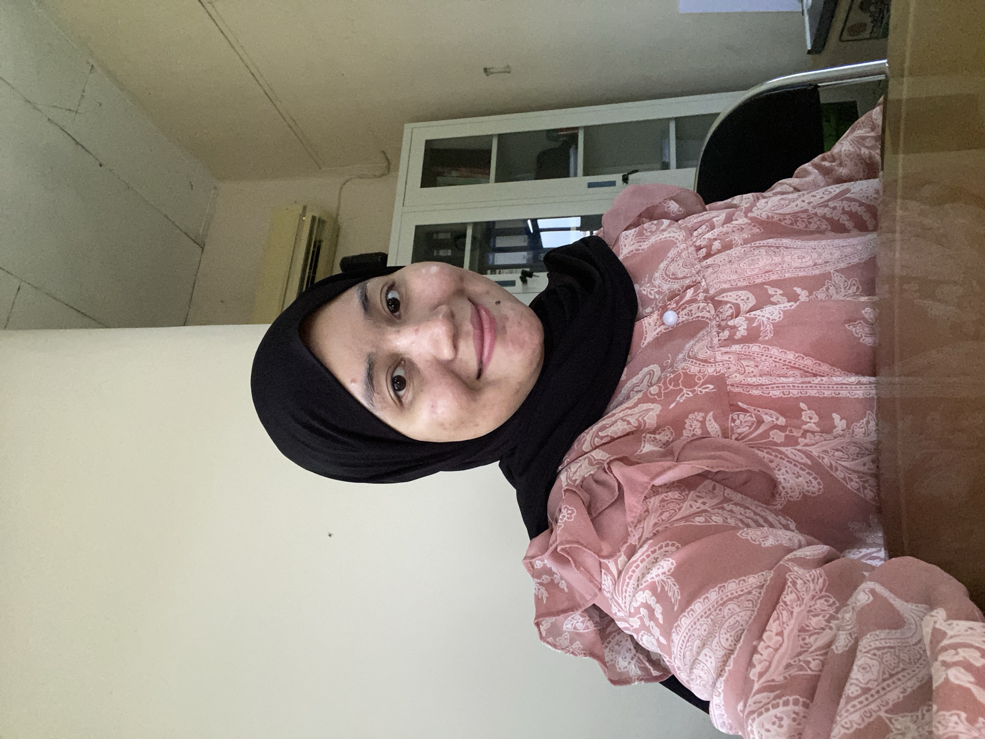 A medium shot captures a woman with fair skin and dark eyes wearing a black hijab and a light pink patterned blouse with ruffled sleeves. She is seated indoors, likely in an office or study, with a neutral-colored wall behind her. A white cabinet with glass doors and shelves containing books and other items is visible to her right. An air conditioning unit is mounted on the wall above the cabinet. The lighting suggests an indoor setting, possibly during daylight hours.