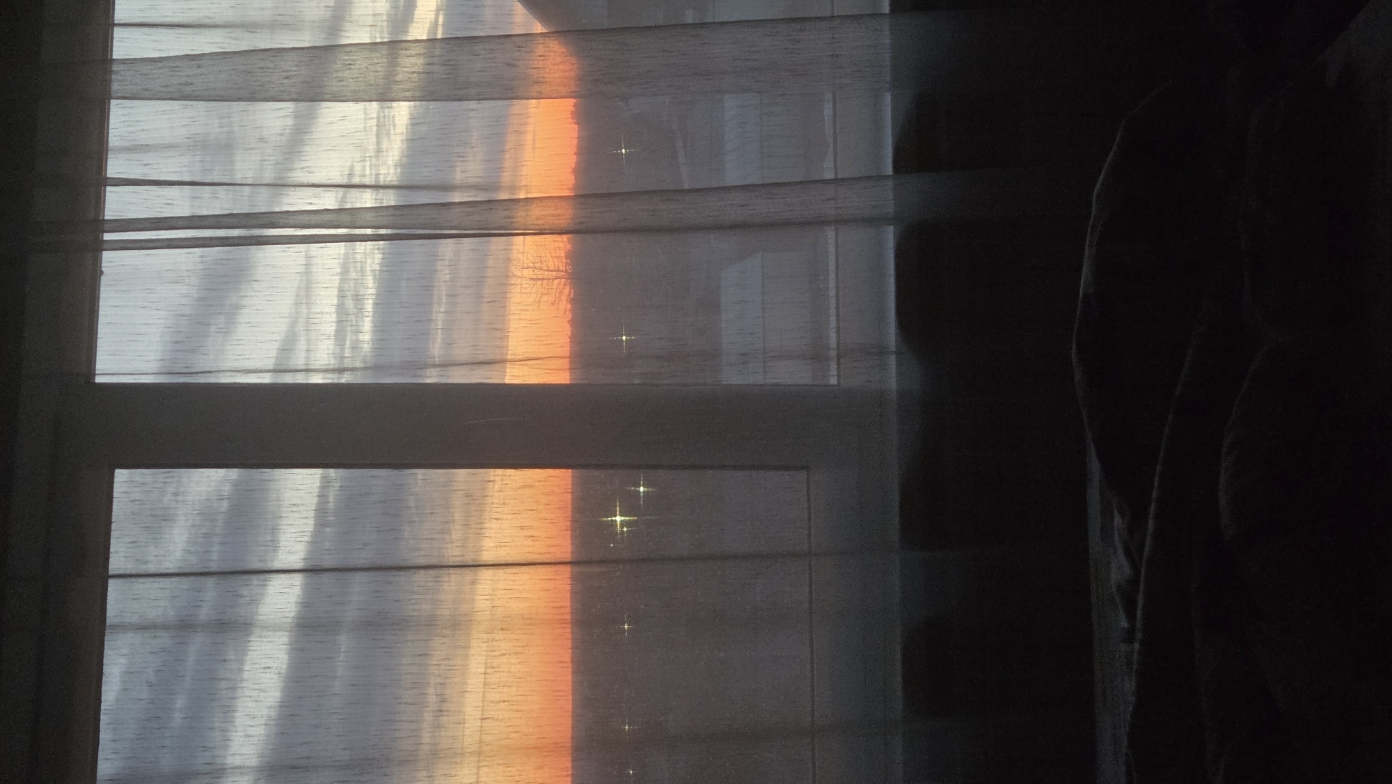 The image depicts a view from inside a room looking out through a window. The window is partially covered with sheer curtains and horizontal blinds. Outside, the scene suggests a sunset or sunrise, with orange and gray hues visible through the window. The time of day seems to be early morning or late evening. A dark, undefined mass, possibly a curtain, is visible on the right side of the image, taking up a significant portion of the frame. The focus of the scene is the interplay of light and shadow through the window coverings. The location in Chaikovsky, Russia, is not discernible from the visual information in the image. No people, actions, or specific objects can be identified beyond the window, curtains, and light.