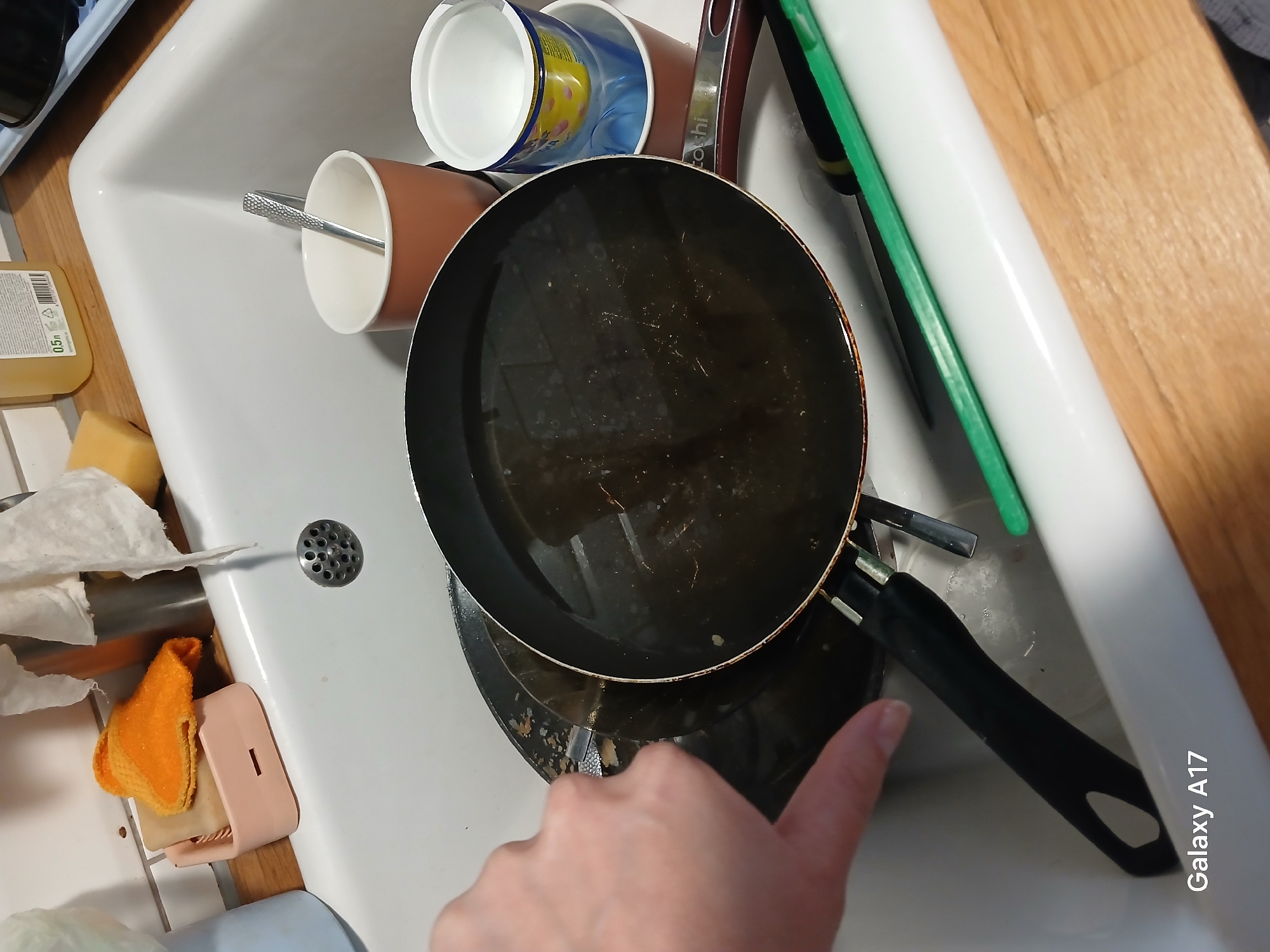 The image displays a cluttered kitchen sink filled with dirty dishes, suggesting a domestic scene in Yekaterinburg, Russia. A light-skinned human hand is visible in the foreground, with an index finger pointing towards the handle of a large, black frying pan.
The white ceramic sink contains several items. A prominent black frying pan, appearing greasy with residue, takes center stage, partially covering a smaller pan beneath it. To the left, a brown and white ceramic mug holds a silver spoon with a textured handle. Above these, a white cup is stacked on a blue cup featuring a yellow polka-dot pattern. A dark red handle with the partially visible text "Toshi" is seen behind these cups.
To the left of the sink, on a wooden countertop, are a yellow dish soap bottle with "0.5 L" visible on its label, a yellow sponge, and crumpled white paper towels. An orange dishcloth and a small pink soap holder are also present. On the right, a green and white kitchen utensil, possibly a scraper or spatula, rests against the sink and countertop. A metallic drain cover is visible in the upper left of the sink.
The scene depicts the aftermath of cooking, with dishes awaiting cleaning. A "Galaxy A17" watermark is present in the bottom right corner of the image.