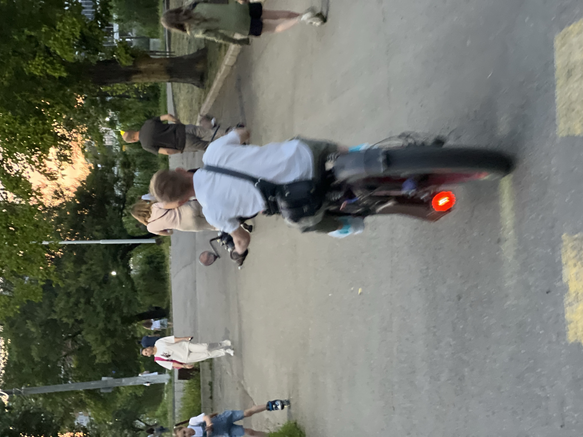 The image depicts a scene in a park or pedestrian area in Krasnodar, Russia. In the foreground, a young person is riding a motorized bicycle with a red taillight illuminated. The rider is wearing a white t-shirt and dark shorts, with a black bag slung across their back. To the left, a young child is on rollerblades, wearing a denim overall dress and a white t-shirt. Further in the background, several other individuals are walking. One woman in a white outfit with a pink backpack is walking towards the left, and another person in a dark shirt and light shorts is walking away from the camera towards the right. A woman in a light pink top and beige pants is also visible, walking away from the camera. In the far right, a young person in a green long-sleeved shirt and dark shorts is walking away. The environment includes paved pathways, trees with green foliage, and glimpses of buildings and power poles in the distance. The lighting suggests it is late afternoon or early evening, with the sun casting a warm glow through the trees.