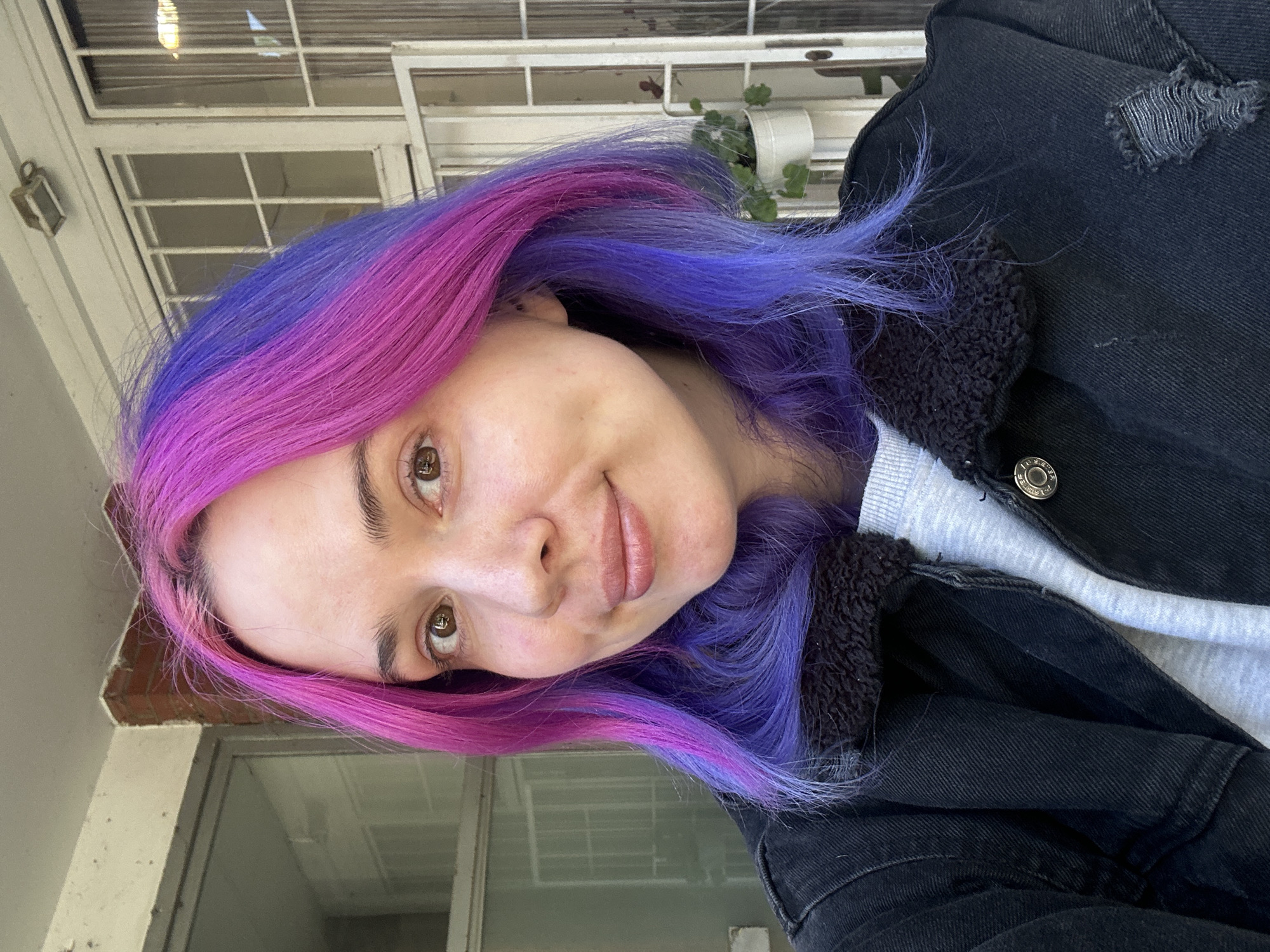 The image displays a young person, likely female, captured in a selfie-style portrait, rotated 90 degrees clockwise. She has distinctive two-toned hair, featuring vibrant magenta or pink on one side and a rich purple on the other, creating a striking split-dye effect. Her brown eyes are open, and she looks directly at the camera with a subtle, gentle smile. Her complexion appears clear and natural.
She is dressed in a black denim jacket, which features a dark, possibly sherpa or fleece, lining visible along the collar and inner lapel. The jacket also shows some distressed or ripped detailing on the shoulder. Beneath the jacket, she wears a light grey sweatshirt. Small metal buttons on the jacket, which appear to have text such as "AMERICAN EAGLE," are visible.
The background suggests an outdoor setting or a brightly lit interior space adjacent to a building. Visible elements include white window frames with multiple panes, a section of a light-colored brick wall, and a small white pot holding green plants, possibly hanging or placed on a ledge. A small, decorative light fixture or lantern is mounted on the wall near the window. The lighting indicates it is daytime, characterized by soft, indirect illumination. The overall impression is relaxed and casual. This scene is located in the City of Zagreb, Croatia.