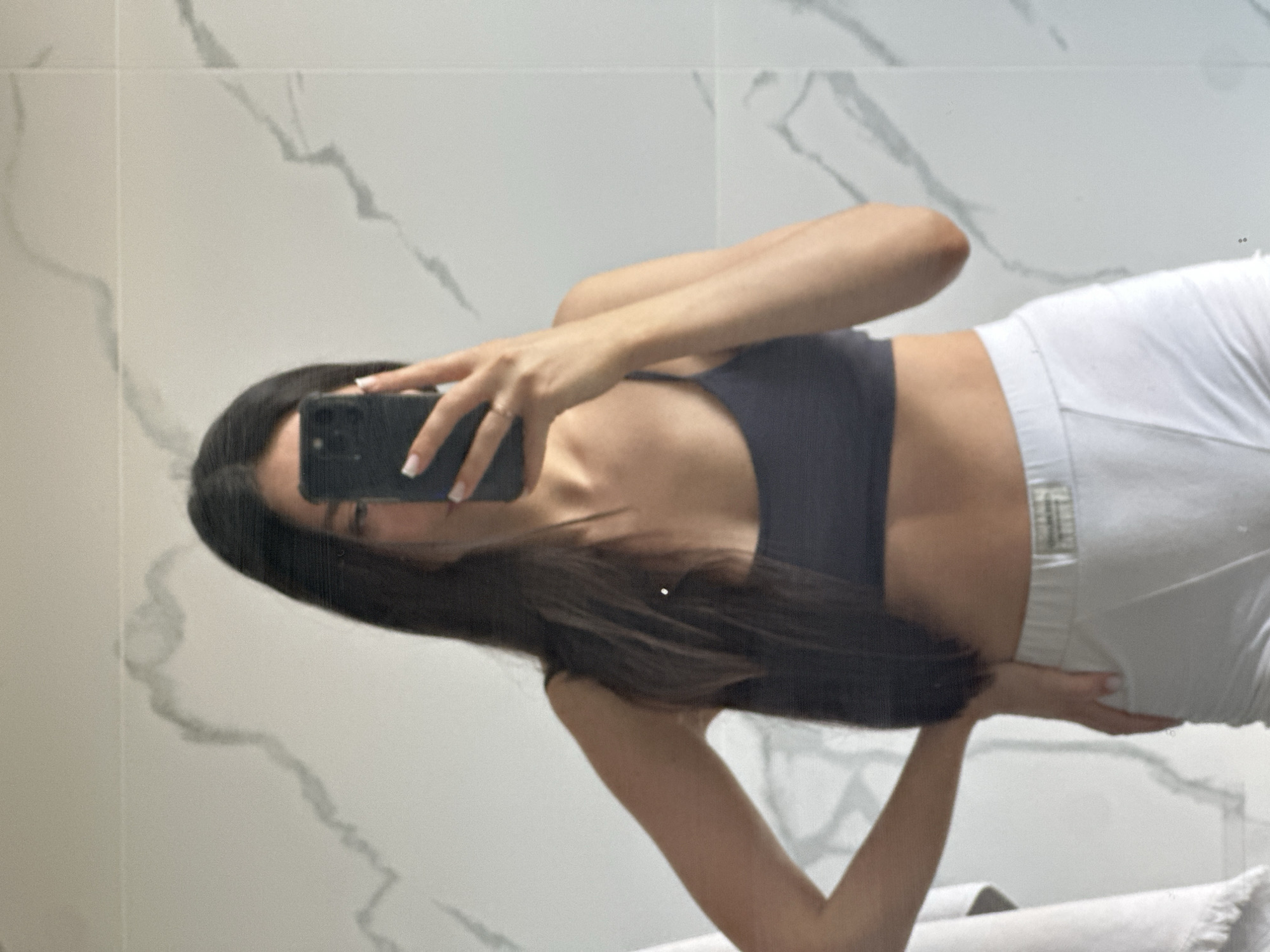A young woman, seen from the waist up, stands in front of a marble-tiled wall, holding a smartphone to take a selfie. She has long, dark hair that falls over her shoulders. She is wearing a dark-colored sports bra and white boxer shorts. Her left hand is on her hip, and her right hand is holding the phone. The background is a tiled wall with a marble pattern. The image is taken indoors, likely in a bathroom. There is no visible indication of the city of Эстачи, Russia.