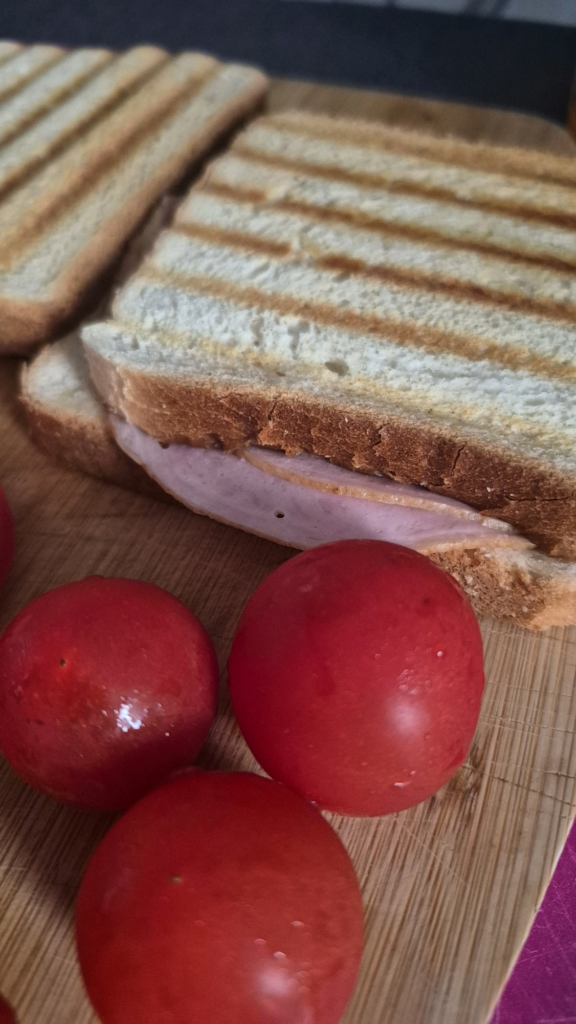 The image shows a close-up, slightly angled view of three bright red cherry tomatoes and a sandwich on a wooden cutting board. The sandwich appears to be made with white bread that has been toasted, creating distinct grill marks. Slices of a pinkish, processed meat, possibly ham or bologna, are visible inside the sandwich. The tomatoes are plump and appear slightly moist. The wooden cutting board has a visible grain pattern. The background is dark and out of focus. There is no indication of the city of Krauja, Latvia, or any specific location within it.