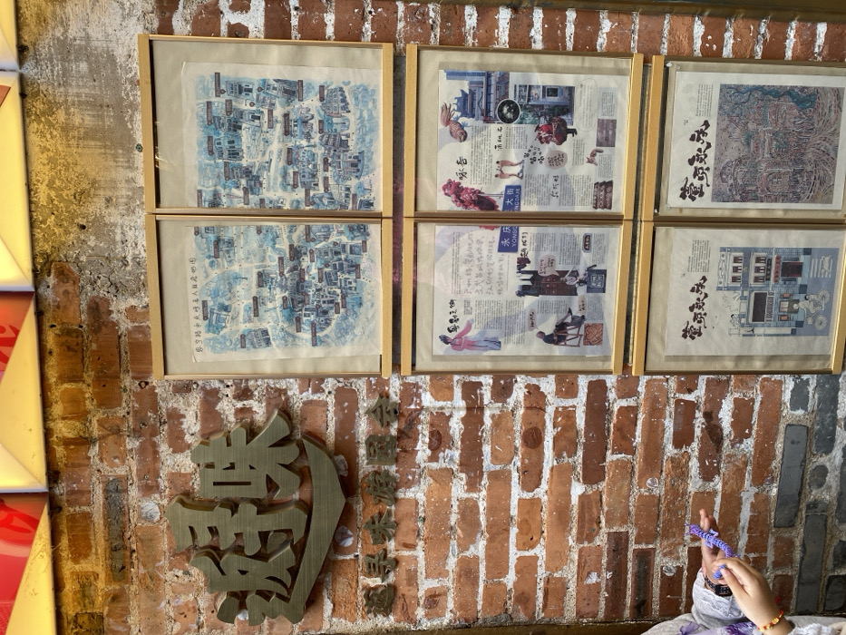 The media file captures an indoor scene dominated by a brick wall adorned with six framed artworks and decorative metal signs. The setting appears to be a cultural space or establishment in Guang Zhou Shi, China.
The upper portion of the brick wall features six rectangular frames arranged in two rows of three. The two frames on the left display illustrative, watercolor-style maps, depicting an intricate layout of buildings and streets. Visible text on these maps includes "您宁路永庆明名人目店地图" (You Ning Road Yong Qing Ming Celebrity Shop Map). The four frames on the right contain a collage of images, illustrations of traditional figures and objects, and extensive Chinese text, reminiscent of historical documents or old newspaper clippings. Phrases like "永庆" (Yong Qing), "YONGC," "大街" (Da Jie), and "ENG DAJE" are discernible, along with "YONG CING GATE" in one instance. Another piece of text, "咏春" (Yong Chun), is also visible, potentially related to one of the framed pieces or the metal signs.
Below the framed artwork, a large, intricately carved or cast metal character in a golden hue is mounted on the brick wall. To its right, a series of smaller, similar metal characters form a phrase. In the bottom right corner of the image, a person's hands are visible, holding a purple, braided object, possibly a decorative string or small toy. A beaded bracelet is also worn on one wrist. The overall impression is one of a richly decorated space, possibly highlighting local history, geography, or cultural heritage. The time of day and weather are not discernible.