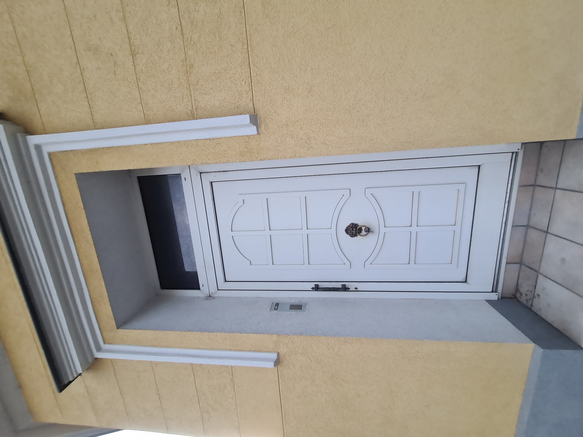 The image depicts a white front door with decorative panels and a lion-head door knocker, set within a light yellow textured wall. The door is framed by white trim that extends upwards to a decorative cornice. Above the door, a dark transom window is visible. To the left of the door, a silver intercom system is mounted on a light grey column. The ground in front of the door is paved with light-colored square tiles. The scene is an exterior view of a building. The image is tagged as being in Vienna, Austria. There are no people or specific actions depicted.