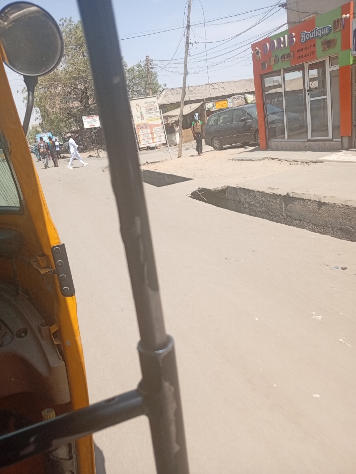 The image depicts a street scene in Sabon Gari East, Nigeria, viewed from the interior of a yellow tricycle (Keke Napep), partially obscured by a black metal structural bar in the foreground. The observer's round side mirror is visible in the upper left.
On the right side of the street, a two-story commercial building with an orange and white facade houses "DORIS Boutique," indicated by prominent signage including phone numbers. The building features large glass doors and windows reflecting the environment. A dark-colored minivan or small SUV is parked curb-side in front of this boutique. Standing on the pavement near the parked vehicle is a male individual wearing a light-colored helmet or headwear and a multi-colored vest, looking towards the left. The pavement on this side exhibits a partially damaged concrete drain or raised curb.
Across the street, on the left, several individuals are walking. One male figure in all white clothing (shirt and trousers) is prominent, crossing from left to right. Further down, multiple utility poles with a dense network of overhead electrical wires are visible. In the background, older, lower-rise buildings with corrugated metal roofs are present, along with large advertising billboards, one of which mentions "CHRIST." A large tree with sparse foliage stands on the left. The road surface appears to be asphalt or packed dirt, with a clear, sunny sky overhead.