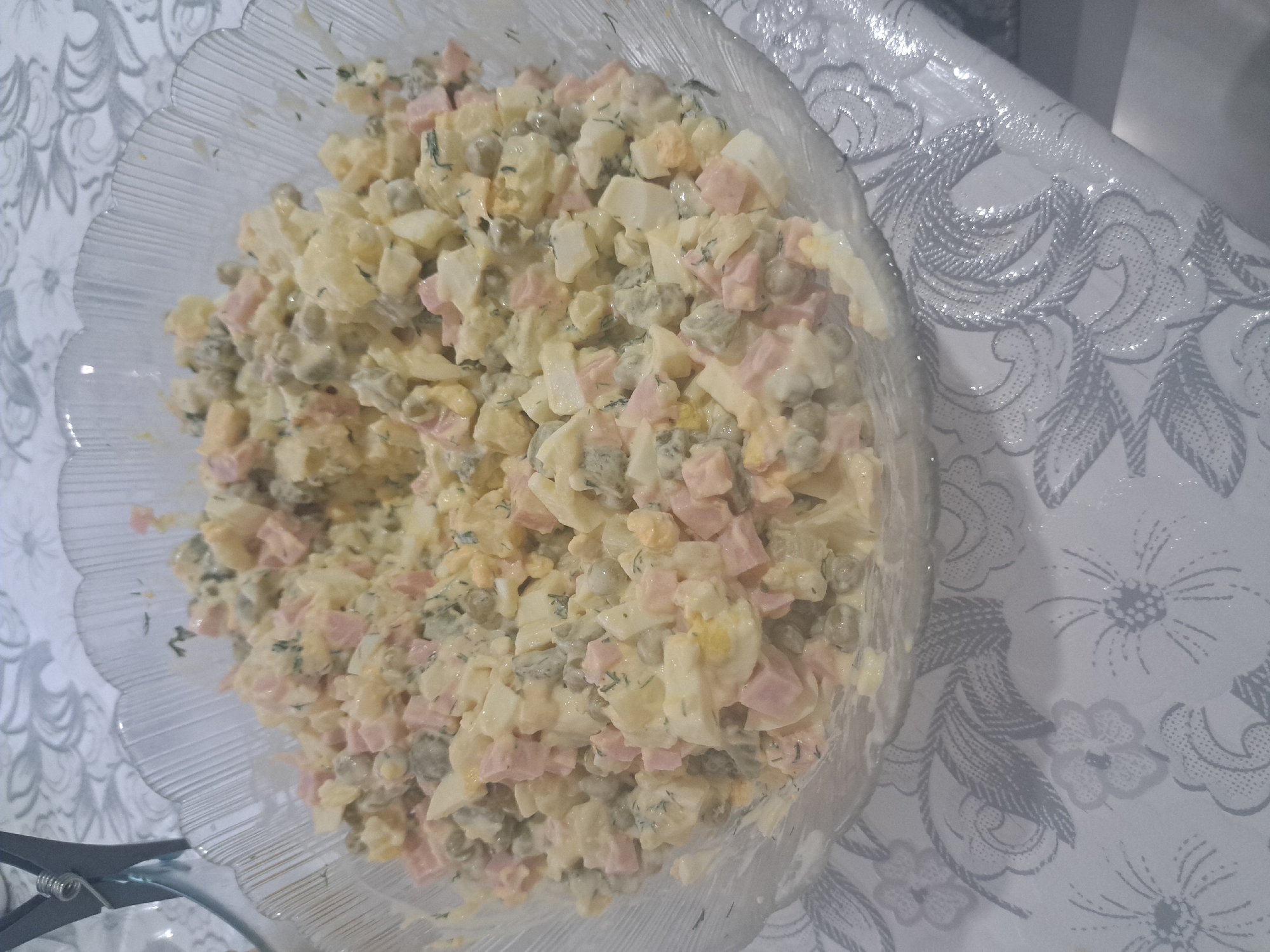 A clear, ribbed glass bowl is filled with a mixed salad. The salad appears to be composed of chopped ham, hard-boiled eggs, peas, and possibly potatoes, all coated in a creamy dressing. Fresh dill is also visible throughout the salad. The bowl sits on a white tablecloth with a grey, floral pattern. In the upper left corner of the image, a part of a grey metal object, possibly a binder clip, is visible. The scene is set indoors, with soft lighting. No specific location in Leningradskaya, Russia can be determined from the visual information provided.