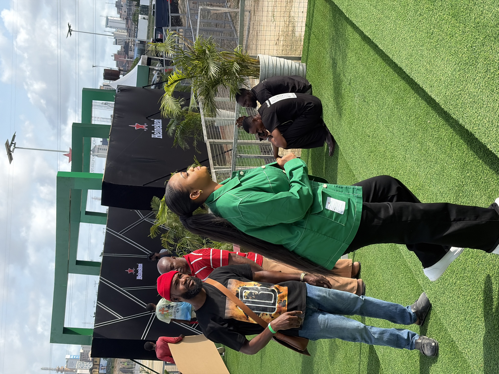 The image shows an outdoor event in Lagos, Nigeria, indicated by the city skyline in the background. The event appears to be sponsored by Heineken, evidenced by the branding on the black backdrop. The foreground features artificial green turf.
There are several people in the image:
A woman with a high ponytail wearing a green jacket and black pants.
A man with a red cap wearing a black t-shirt, jeans, and a brown shoulder bag.
A man in a red and white striped shirt.
Two men are crouching down with their backs facing.
Several other people are visible but less distinct. A possible stage structure is behind the crowd. A palm tree is placed in a large pot on the turf.