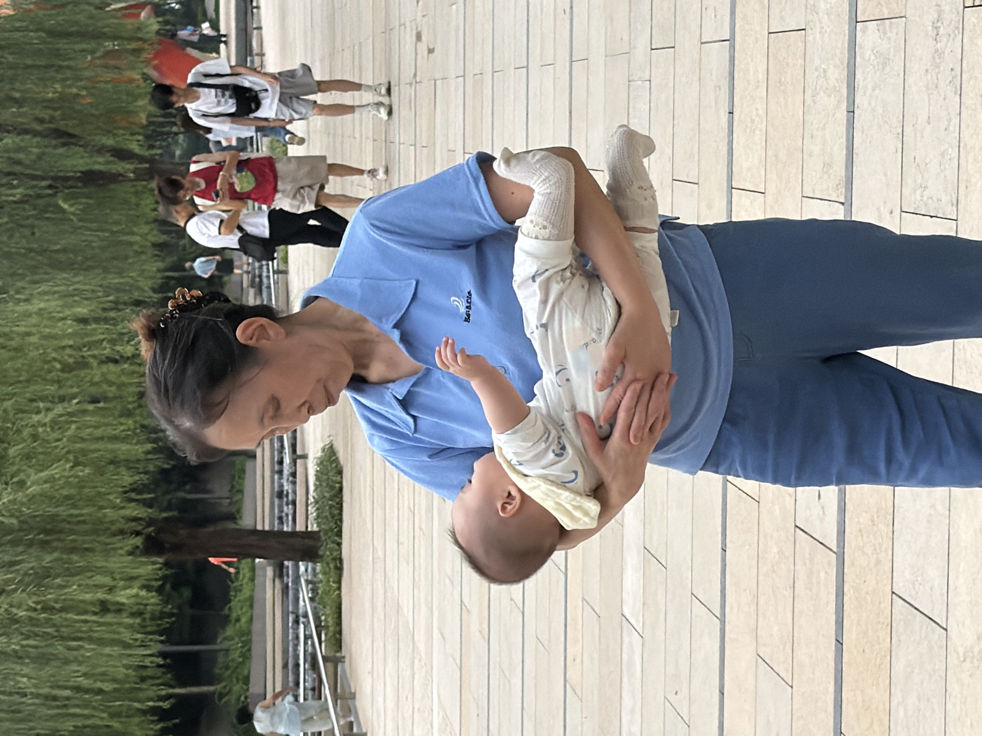 A woman in a blue polo shirt and blue pants is holding a baby. The woman has dark hair pulled back with a clip. The baby is wearing a white onesie and white socks. In the background, there are trees and other people walking. The scene appears to be an outdoor public space with paved walkways. The location is identified as Yuzhong District, China.