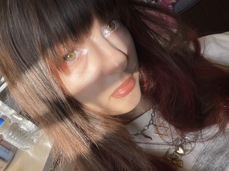 A close-up, angled portrait captures a female subject indoors, described as being in Petah Tikva, Israel. The subject has dark brown hair with visible reddish highlights, parted to the side, and bangs partially obscuring her forehead. Her face, tilted upward and to the right, is illuminated by direct sunlight, casting shadows across her nose and chin. She possesses fair skin, light green or hazel eyes with visible white eyeliner on the lower waterline, and her lips are covered with a peach-toned lip gloss. She wears a light-colored, possibly white, collared garment. Adorning her neck are multiple silver-toned necklaces; one features a prominent Star of David pendant, another a red chili pepper (cornicello) charm, and additional smaller pendants including a heart and an indistinct winged figure. The background is blurred, showing an indoor environment with several clear plastic bottles, likely containing water, and other containers visible on a surface, suggesting a kitchen or utilitarian area.