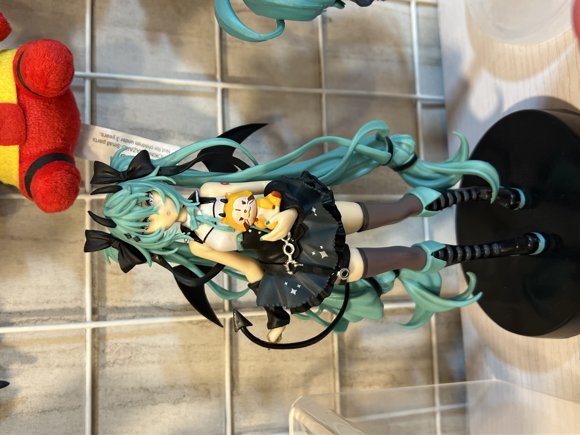 A close-up, eye-level shot features a mint-green-haired anime figurine with black devil horns and bat-like wings. The figurine is wearing a black outfit with a short, ruffled skirt adorned with white star-like patterns and silver accents. She holds a small, orange and white cat-like plush toy in her arms. The figurine stands on a black circular base. The background is a textured grid of light-colored material, possibly a wall or display panel. A white label with black text is visible behind the figurine's head, indicating "CHOKING HAZARD - Small parts. Not for children under 3 years." The overall lighting is soft, with some shadows suggesting overhead illumination. There is no discernible urban environment or location in Arica, Chile.