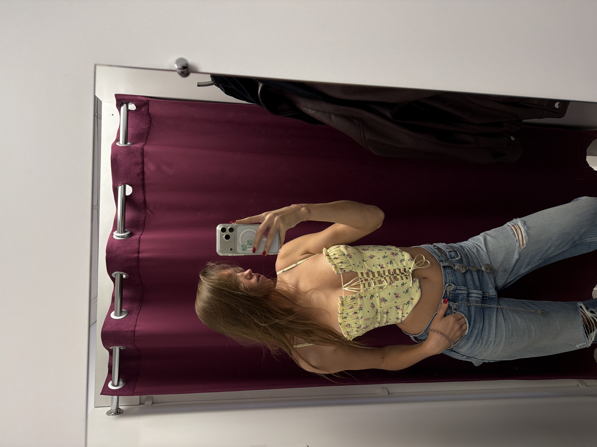 A young woman is taking a mirror selfie in a dressing room. She is wearing ripped light blue jeans, a yellow floral top, and a chain belt. A dark garment is hanging on a hook above the burgundy curtain that serves as the backdrop. The dressing room appears to be located in a retail establishment; the location in the city of Nakhodka, Russia, cannot be determined from the image.