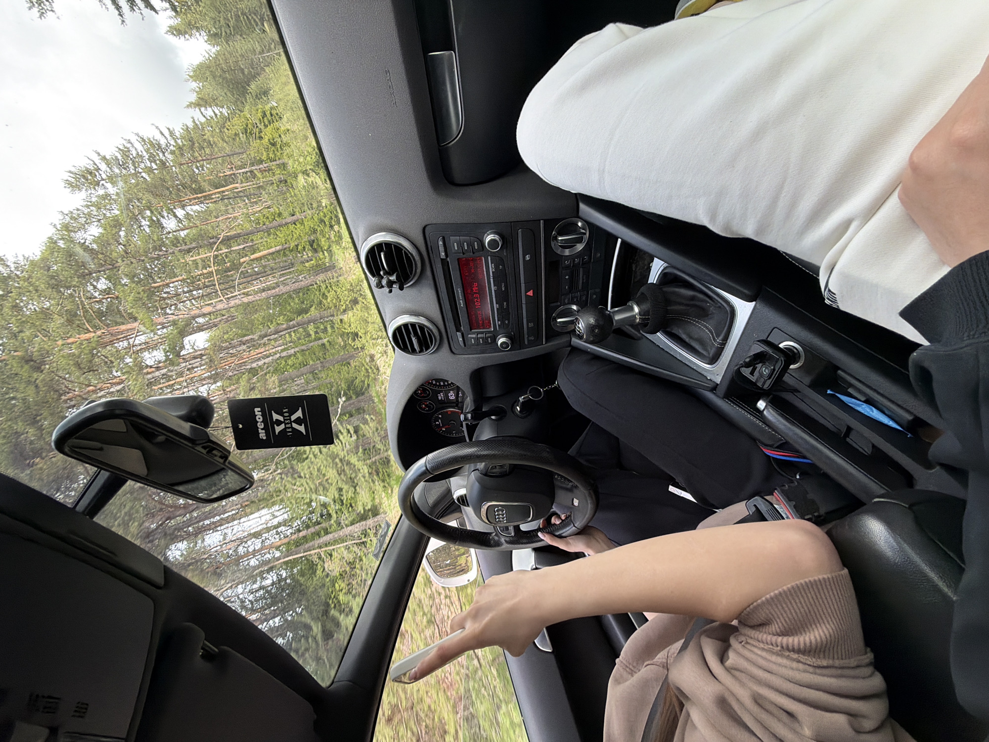 The interior of a car is shown from the driver's perspective, with trees visible through the windows. A person is in the driver's seat, holding a phone in their left hand. Another person, wearing white pants, is in the passenger seat. An "Areon Version" air freshener hangs from the rearview mirror. The radio displays "Audi concert 102.5 MHz." No location information is provided regarding the city of Kasak, Bulgaria.