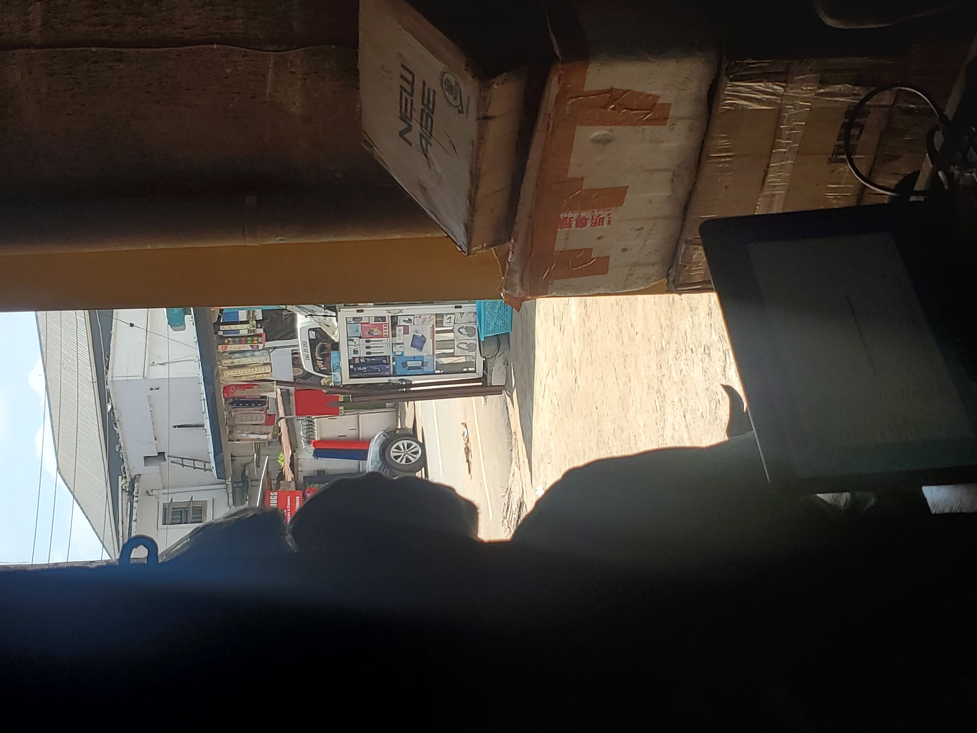 The image depicts a scene observed from inside a shaded structure, looking out onto a sunlit commercial street in Fegge, Nigeria. In the immediate foreground, a person is silhouetted, facing away from the camera and towards a laptop with a glowing screen. The individual appears to have short, dark hair and subtle facial hair visible on the jawline. To the right of the frame, several cardboard boxes are stacked. The top box is light brown with "NEW AGE" printed in dark letters, accompanied by an illegible logo. Lower boxes are taped with brown parcel tape, one showing partial Chinese characters.
Outside the structure, a busy street scene unfolds. The ground is a light-colored, unpaved or dirt surface. A black car is visible on the street. To the left, across the street, a white building with a grey corrugated roof dominates the background. Adjacent to it, another white building features a wooden-framed window and a wooden ladder leaning against its facade. Along the street, market stalls or vendor displays are visible. One partially legible sign reads "RUGS" and "Project & Flowers." A mobile display stand, possibly for electronics, is positioned on the right side of the street, showing various small items and a blue plastic basket at its base. A white van with "UWA ON AIR" text on its side is partially visible behind this display. The overall environment suggests a bustling urban commercial district.