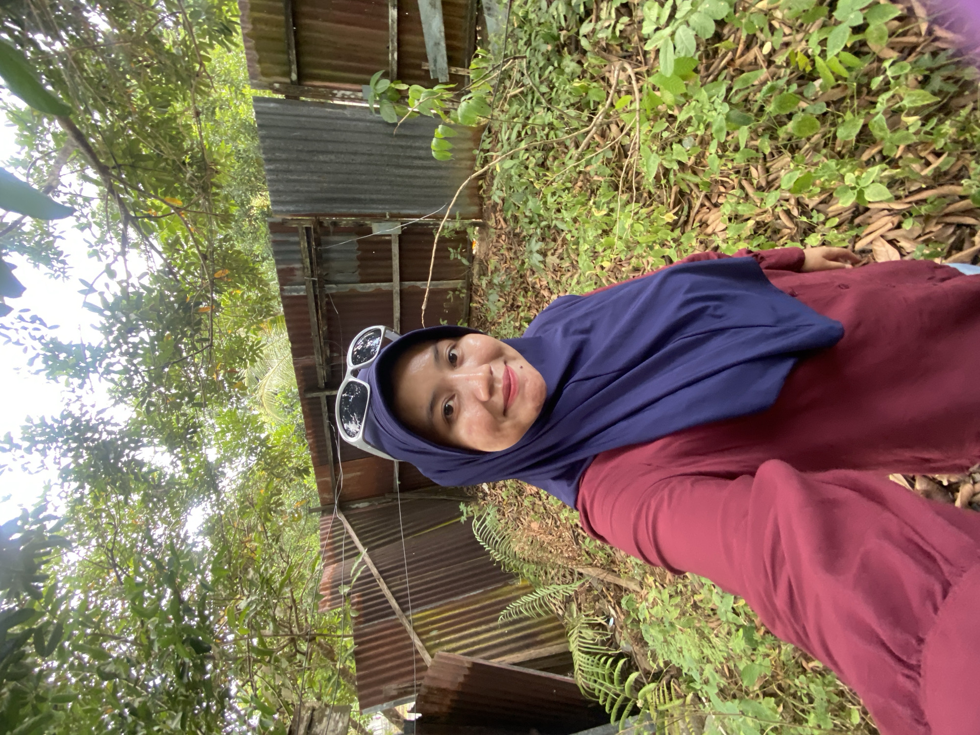 A woman wearing a dark blue hijab and a maroon shirt is taking a selfie outdoors. She has sunglasses perched on her head and is smiling at the camera. Behind her, a rusty, corrugated metal fence separates the area from dense foliage and trees. The ground is covered in leaves and small plants, suggesting a somewhat overgrown or natural environment. The overall setting appears to be a residential or undeveloped area within Banjarbaru, Indonesia.
