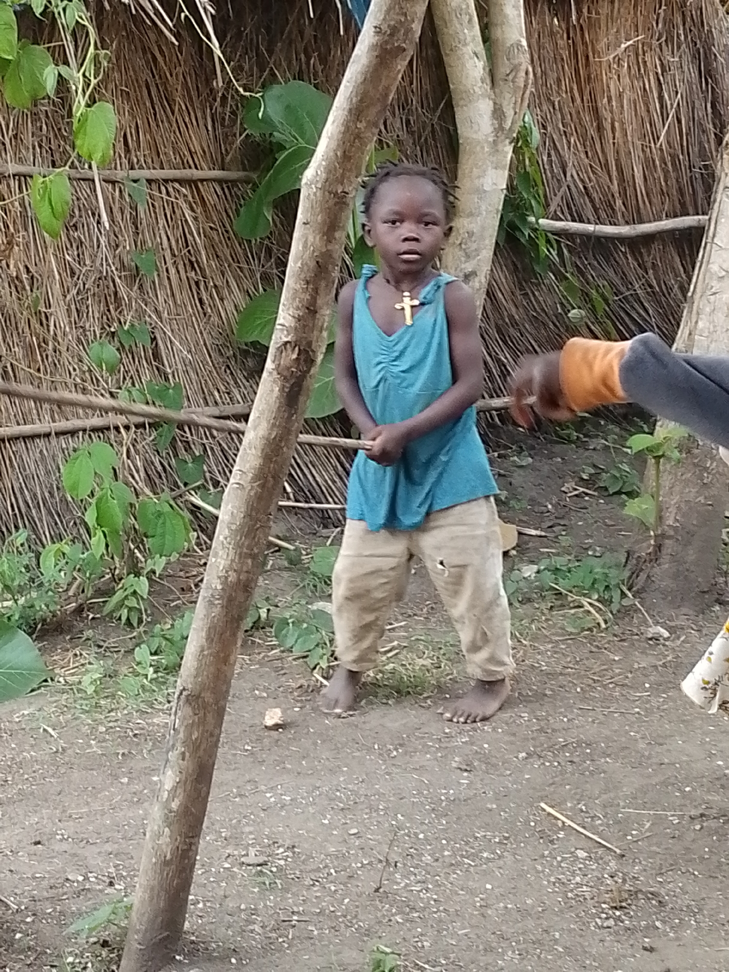 A young Black child, appearing to be between four and six years old, stands in front of a thatched wall and two trees in Otulu, Nigeria. The child is wearing a teal-colored tank top with ruching at the neckline and khaki pants that are ripped at one knee. A gold-colored cross necklace hangs around the child's neck. The child is barefoot. To the right of the child, an adult's arm, wearing an orange sleeve and grey cuff, is extended into the frame, as if interacting with the child or something out of frame. The ground is dirt with some scattered pebbles and dry vegetation. The overall impression is one of a simple, outdoor setting in a rural or semi-rural environment.