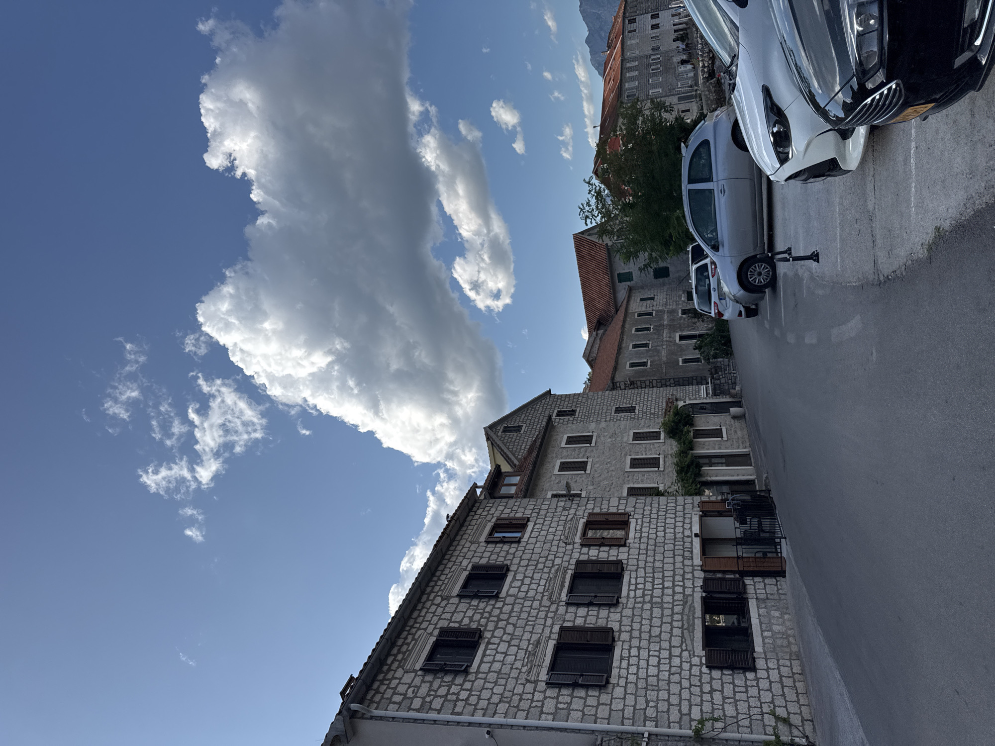 A street scene in Dobrota, Montenegro, features traditional stone buildings with dark shutters and red-tiled roofs lining a paved road. Several parked cars are visible, including a silver hatchback, a white sedan, and a black car. The sky is a bright blue with scattered white cumulus clouds. In the background, a range of hills is visible. The overall impression is one of a quiet, picturesque coastal town.