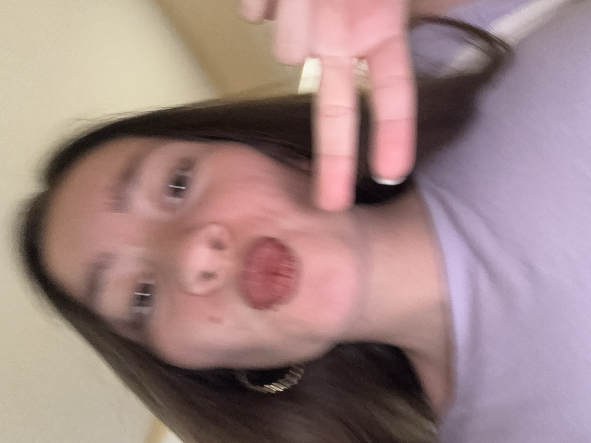 The image is a close-up, out-of-focus selfie of a young person with dark hair, wearing a light purple t-shirt and a hoop earring. Their mouth is slightly open, and they are holding up two fingers in a peace sign. The background is blurred and appears to be an indoor setting with a light-colored wall. The overall impression is casual and informal.
