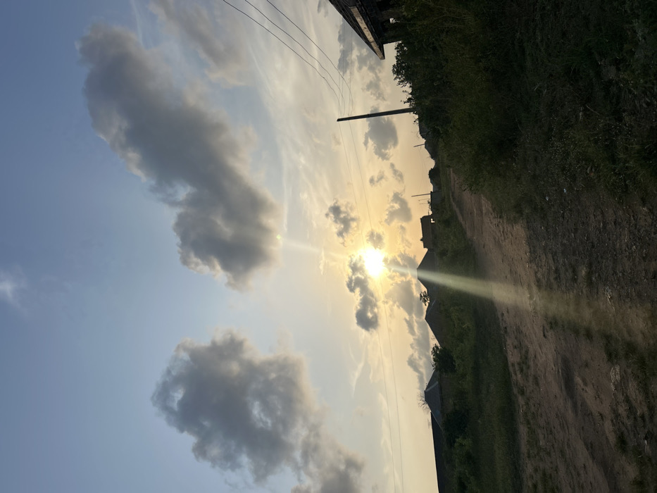 The image depicts a semi-rural or residential area within Debasco, Nigeria, at either sunrise or sunset. The sky displays a gradient of light blue at the zenith transitioning to a warm yellow-orange hue near the horizon, with several scattered cumulus clouds. The sun, a bright luminous disc, is positioned slightly left of center, low on the horizon, emitting distinct rays of light that extend downwards and diagonally to the right.
In the foreground, an unpaved dirt path, approximately 2-3 meters wide, curves gently upwards towards the center-right of the frame. The path's surface appears uneven. Dense green vegetation, composed of various grasses and bushes, flanks both sides of this path, with a more significant concentration along the right edge.
In the mid-ground, several low-rise structures with rounded or sloped roofs are silhouetted against the bright horizon, likely residential buildings. On the far right, a dark utility pole stands vertically, supporting multiple overhead power lines that traverse horizontally across the upper-mid portion of the image. Adjacent to the utility pole, a partial view of a building structure, possibly under construction or in disrepair, is visible on the far right. No individuals, vehicles, or animals are present in the scene.