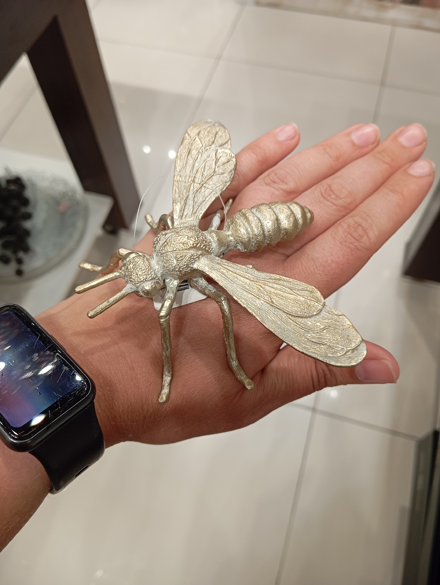 A decorative, gold-colored insect figurine rests on an open palm and fingers of a person wearing a black smartwatch on their wrist. The insect has a segmented body, two pairs of veined wings, and six legs. The background consists of a tiled floor and a blurred display case with dark objects. The scene appears to be indoors, possibly in a retail environment. The location is not specified to be in the city of Orenburg, Russia.