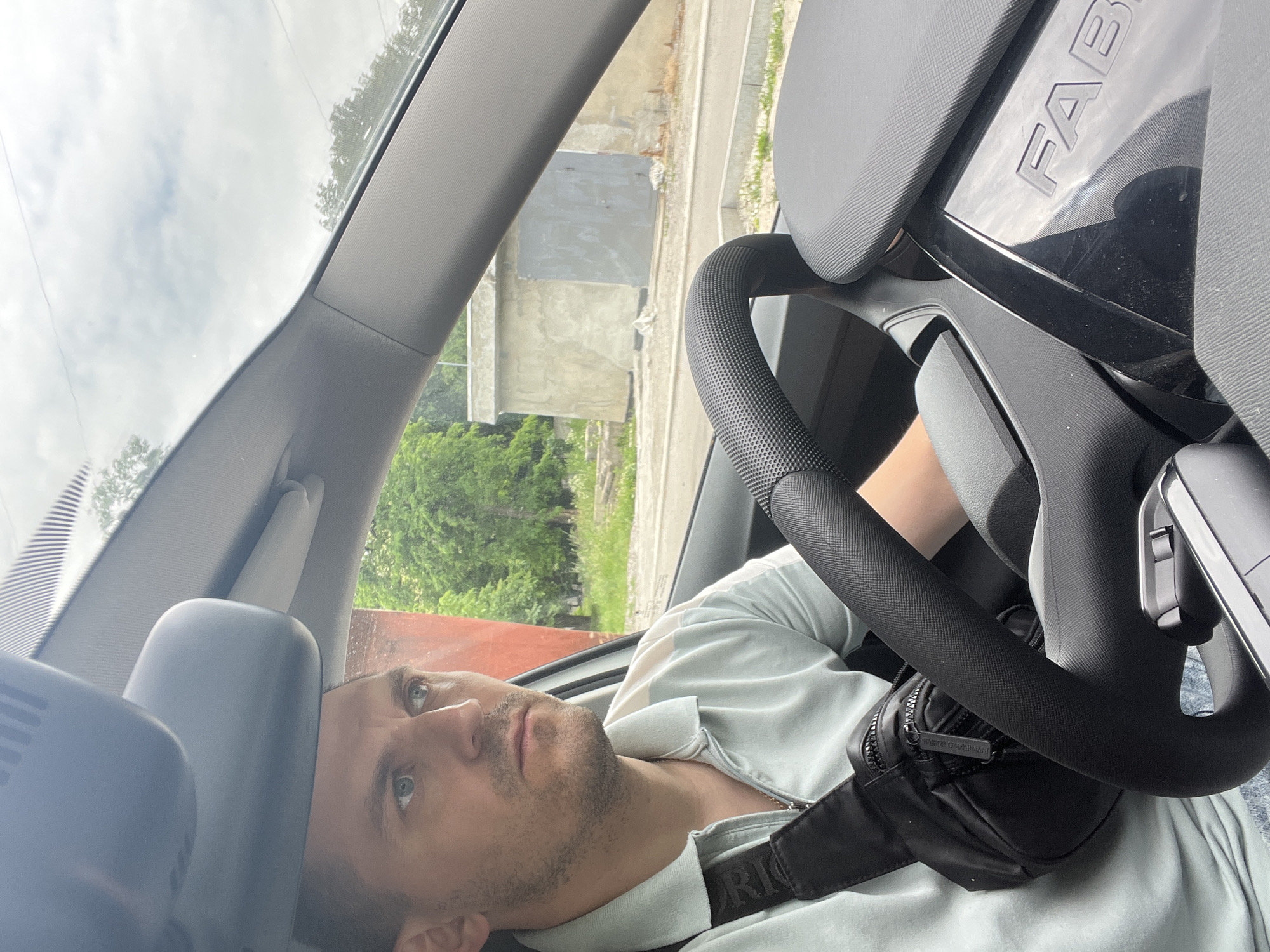 A man is sitting in the driver's seat of a car, looking to the left. He is wearing a light blue polo shirt and a black fanny pack across his chest. The interior of the car is visible, including the steering wheel and a portion of the dashboard. Through the windshield, an outdoor scene is visible with trees, a building, and a cloudy sky. Based on the context of the provided location, the scene is in Kharkiv, Ukraine.