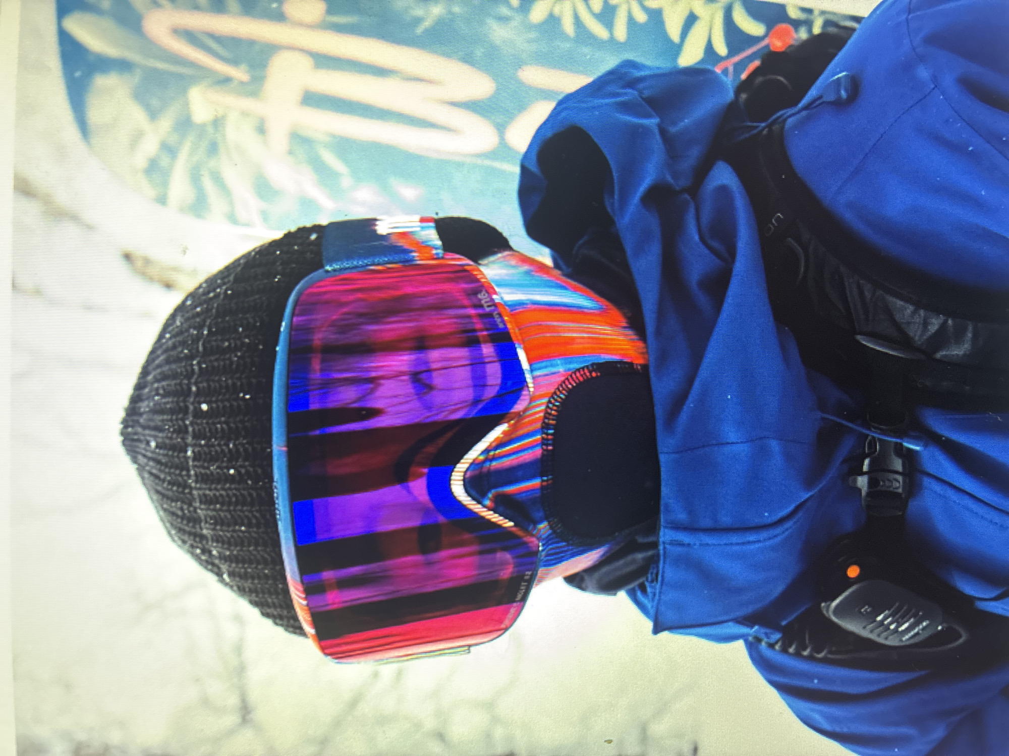 A close-up, profile shot of a person engaged in winter sports, seen from the upper body. The image, initially oriented sideways, features noticeable horizontal scan lines, suggesting it is a photo of a digital screen.
The individual wears a black knit beanie, lightly dusted with what appears to be snow or fine particles. Their face is largely obscured by prominent, highly reflective ski goggles, which dominate the central focus. The goggle lens displays a strikingly vibrant, multi-colored pattern with vertical streaks of purple, blue, red, and orange, creating a dynamic and abstract effect. Visible text on the goggle strap and lower left lens includes "Anon," "M4," and "SONAR," indicating the brand and model.
Below the goggles, a dark, possibly black, neck warmer or balaclava is visible. The person is clad in a bright blue winter jacket, also flecked with small white specks. Over the jacket on the right side, a black strap from a backpack or vest is visible, secured by a black buckle. Attached to this strap is a small black device, possibly a radio or avalanche beacon, which features a textured grille and a glowing orange indicator light. Faint, partially visible text that reads "on" can be seen on the black strap of the backpack or vest.
The background is softly blurred, showing a light blue area with abstract yellow and orange shapes, reminiscent of stylized foliage or graffiti. This could be part of a snowboard's graphic design or a painted backdrop, suggesting an outdoor, snowy environment during daytime. The overall scene captures a moment of readiness for winter recreation. While no specific location cues are present within the image, the provided context indicates the location is Asenovgrad, Bulgaria.