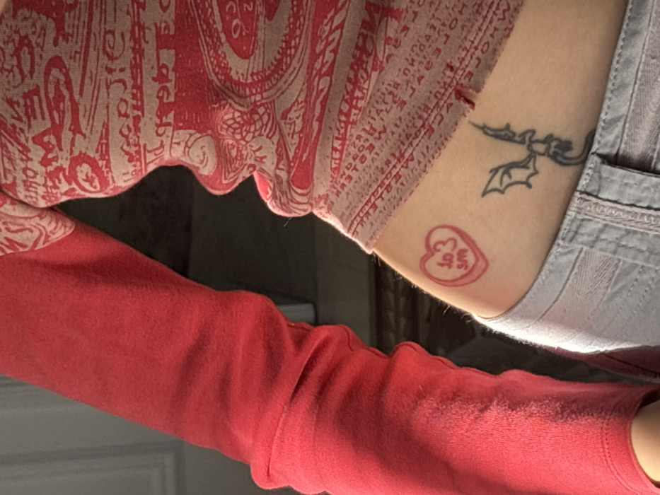 The image presents a close-up, waist-level view of a person, located in Barcelona, Spain. The subject's left arm is visible, clad in a long-sleeved, solid red garment. The torso is covered by a form-fitting top featuring a red and beige pattern of text or stylized graphics, with a ruched or ruffled vertical seam. The subject's midriff and upper hip are exposed. On the left hip, there are two distinct tattoos: a red outline of a heart, resembling a "conversation heart" candy, containing the partially legible text "TO 3 SM"; and below it, a black outline tattoo of a winged creature, consistent with a dragon or gargoyle, positioned on the lower back/hip area. The subject is wearing light gray denim jeans or trousers, with the waistband and upper thigh visible. The background is blurred, dark, and indistinct, indicating an indoor environment with low or indirect lighting. No specific actions or interactions are taking place.
