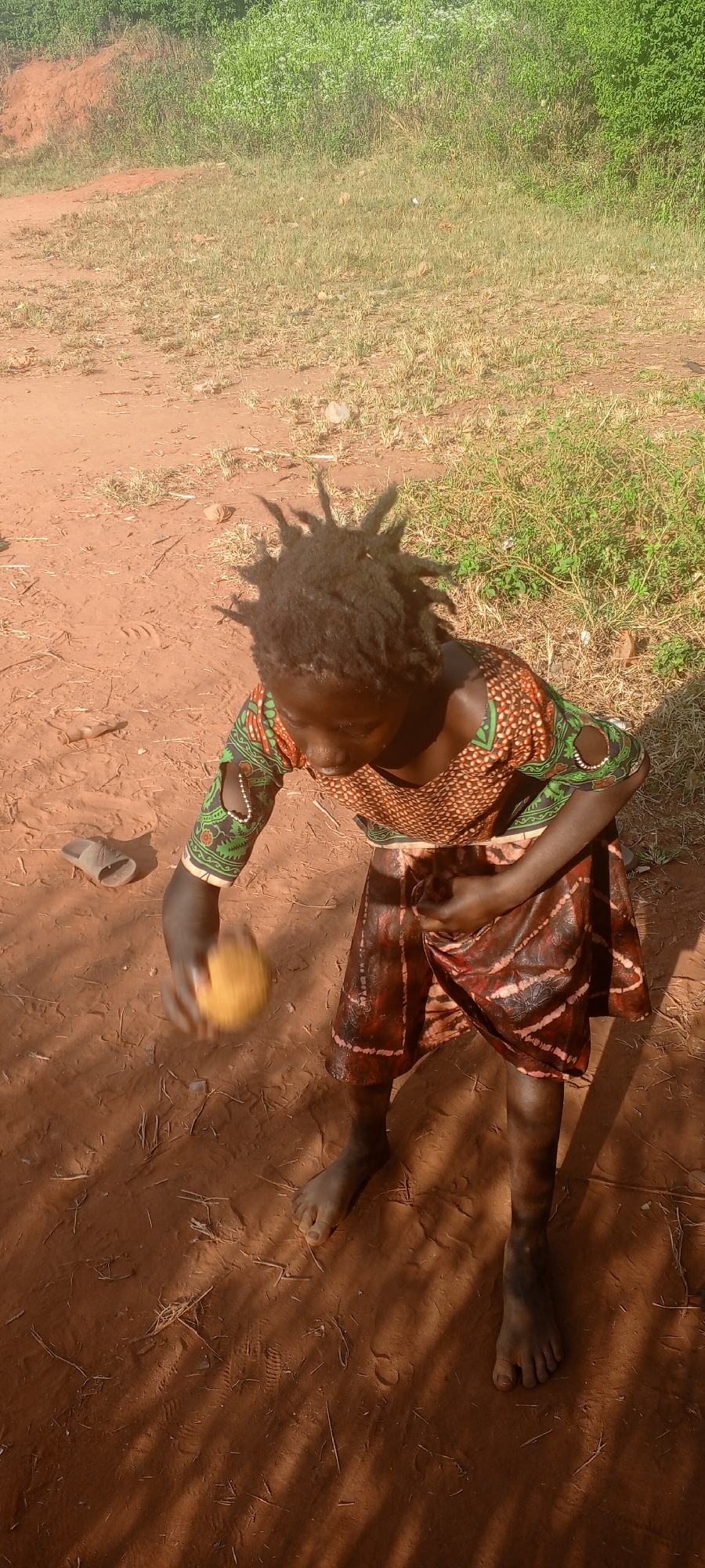 In Ngalula, Democratic Republic of the Congo, a barefoot child with dreadlocks stands on reddish-brown soil. They are wearing a patterned dress with green and brown hues. The child holds a yellow object in their outstretched hand. A lone sandal rests on the ground nearby. Vegetation is visible in the background. Shadows cast across the ground suggest strong sunlight.