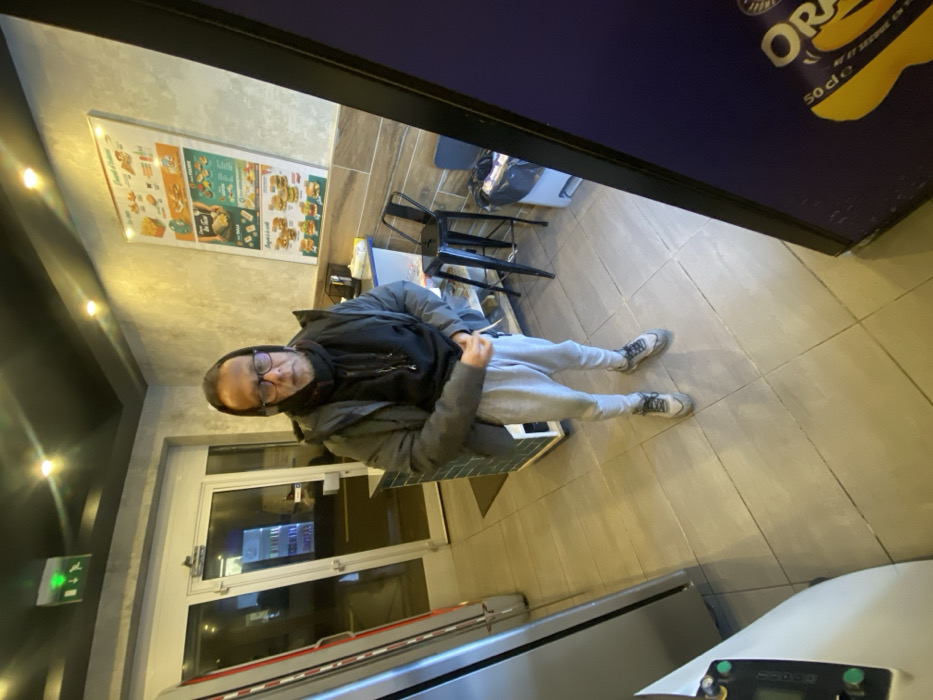 A man wearing glasses, a black hoodie, a gray jacket, and gray sweatpants sits at a table in what appears to be a restaurant. He is looking towards the camera. Behind him are tables and chairs, and a menu board on the wall. A door is on the left side of the image. The shot is taken from a high angle. The frame is partially obstructed by a cooler, obscuring parts of the view.