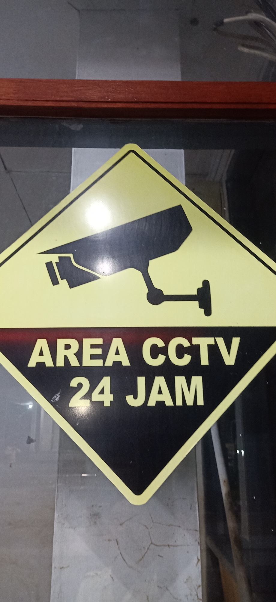 The image shows a diamond-shaped yellow and black sign with a silhouette of a CCTV camera on the top half and text on the bottom half. The text reads "AREA CCTV 24 JAM". The sign is mounted on a concrete pillar and appears to be indoors, possibly in a building or public space. The background is blurred, but it seems to show some reflective surfaces and possibly some wires hanging. The sign indicates that the area is under CCTV surveillance 24 hours a day. The image does not provide any information about the specific location within Tambontunong, Indonesia.