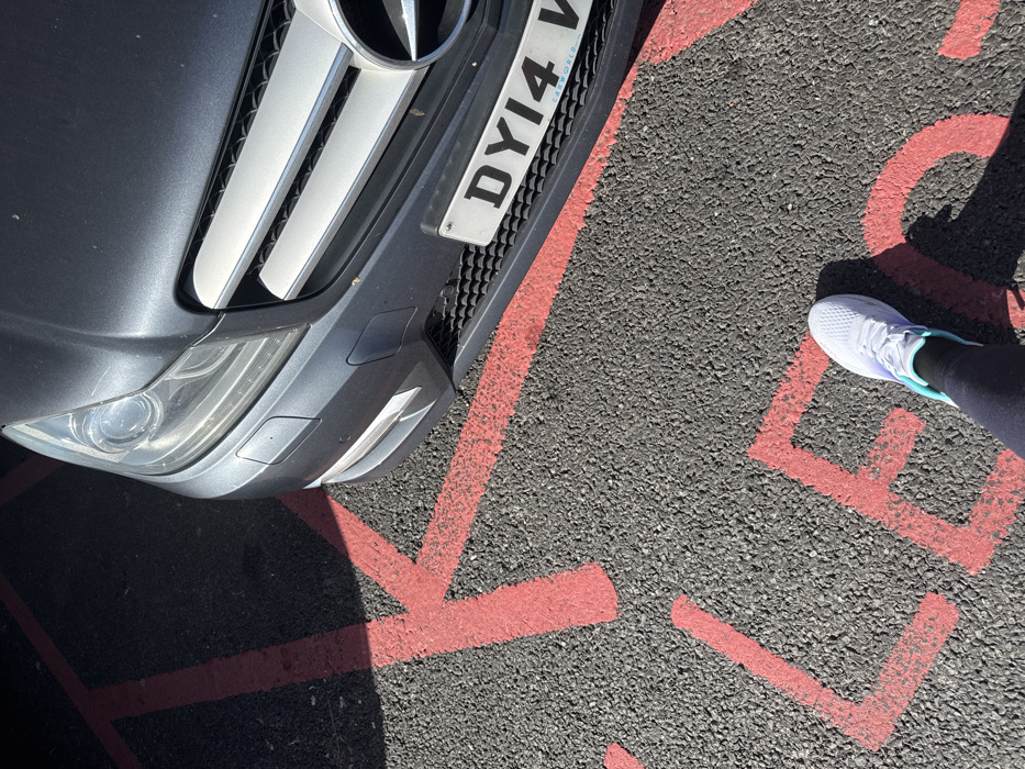 A ground-level, partial view captures the front section of a dark grey Mercedes-Benz sedan positioned on asphalt pavement. The vehicle displays a UK registration plate 'DY14 V' mounted within a frame branded 'CARWORLD'. Red-painted markings are visible on the asphalt surface; these include a distinct 'K' in the upper left, and partial letters 'L' and 'E' in the lower-center. A person's left foot, wearing a white athletic shoe with prominent turquoise accents and black leggings, is visible in the lower right foreground. The scene is illuminated by strong sunlight, casting shadows. Based on the license plate frame, the location is identified as a car park or dealership associated with Carworld UK Ltd on Northfield Avenue, Kettering, United Kingdom.