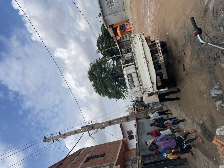This outdoor street scene in Luanda, Angola, depicts a busy, unpaved road under an overcast sky with patches of blue. The ground is rough, with visible dirt and muddy puddles. The time of day appears to be daytime, possibly mid-morning or afternoon.
In the midground, a white flatbed truck is parked facing away from the viewer, its rear visible. The truck has a license plate that reads "LD 597 HP" and the number "88" marked on its side. To the right of the truck, a portion of a motorcycle handlebar can be seen. Behind the truck, a large tree with dense green foliage stands out, and some small market stalls or kiosks are visible.
Several men are gathered on the street. One man in a light-colored jacket and dark pants is standing near the back of the truck. Closer to the foreground, a group of men in casual attire, including t-shirts, shorts, and one in a purple hoodie, are standing and congregating. Some appear to be looking towards the camera, while others are interacting among themselves. On the far right foreground, what appears to be a basket containing eggs or similar produce is visible.
To the left, a utility pole with multiple power lines extends across the frame, leading towards a red brick building under construction. The scene captures a moment of daily life in a bustling, developing urban area, with people gathered on the street amidst vehicles and local architecture.
