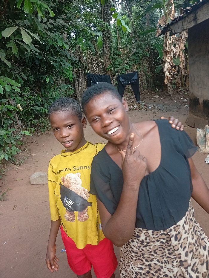 Two individuals are positioned outdoors in Eziudo, Nigeria. On the left stands a male child, wearing a yellow long-sleeved shirt with a brown teddy bear graphic and red shorts. He has short dark hair and a neutral facial expression, looking forward. To his right, an adult female with short dark hair wears a black v-neck top and a leopard-print skirt. She smiles broadly towards the camera, her left index finger raised and bent, and her right hand resting on the child's left shoulder. The ground surface is reddish-brown earth. The background features dense green tropical foliage. To the far right, a rudimentary structure is partially visible, with dried, large plant leaves, consistent with banana leaves, attached or leaning against it. Two pairs of dark trousers are hanging on a visible clothesline in the mid-ground, suspended between trees. A light-colored stone is present on the ground to the left of the child.