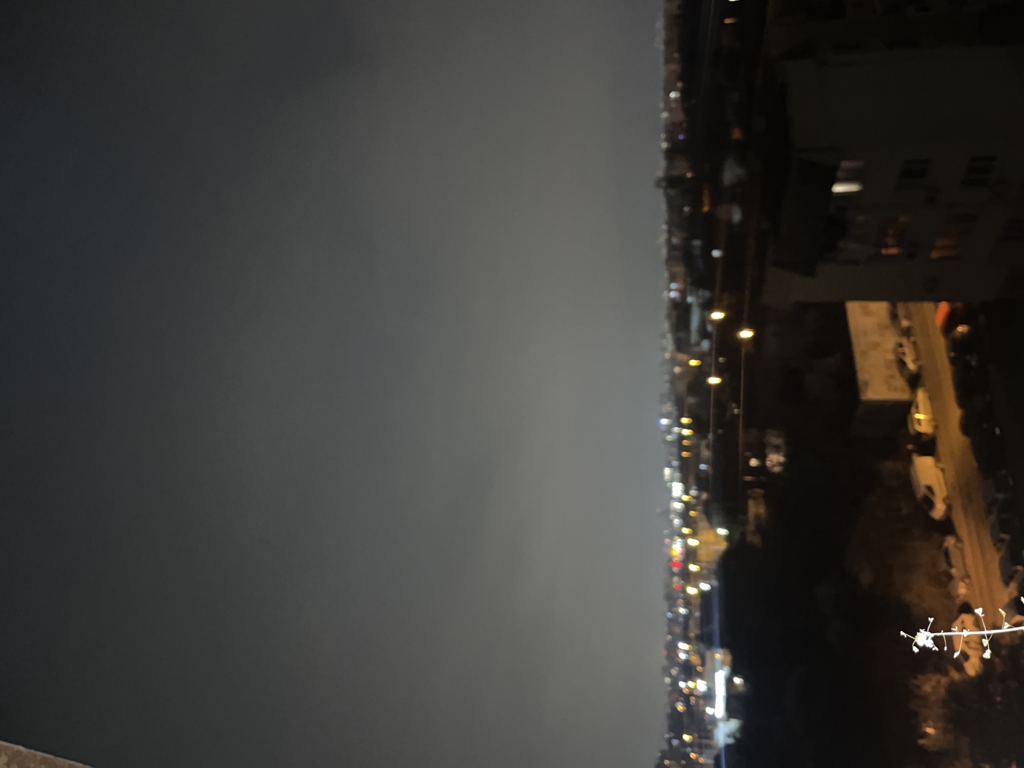 The image shows an aerial night view of Sofia, Bulgaria. Streetlights line a road with parked cars. A building is visible to the right. The sky is overcast.